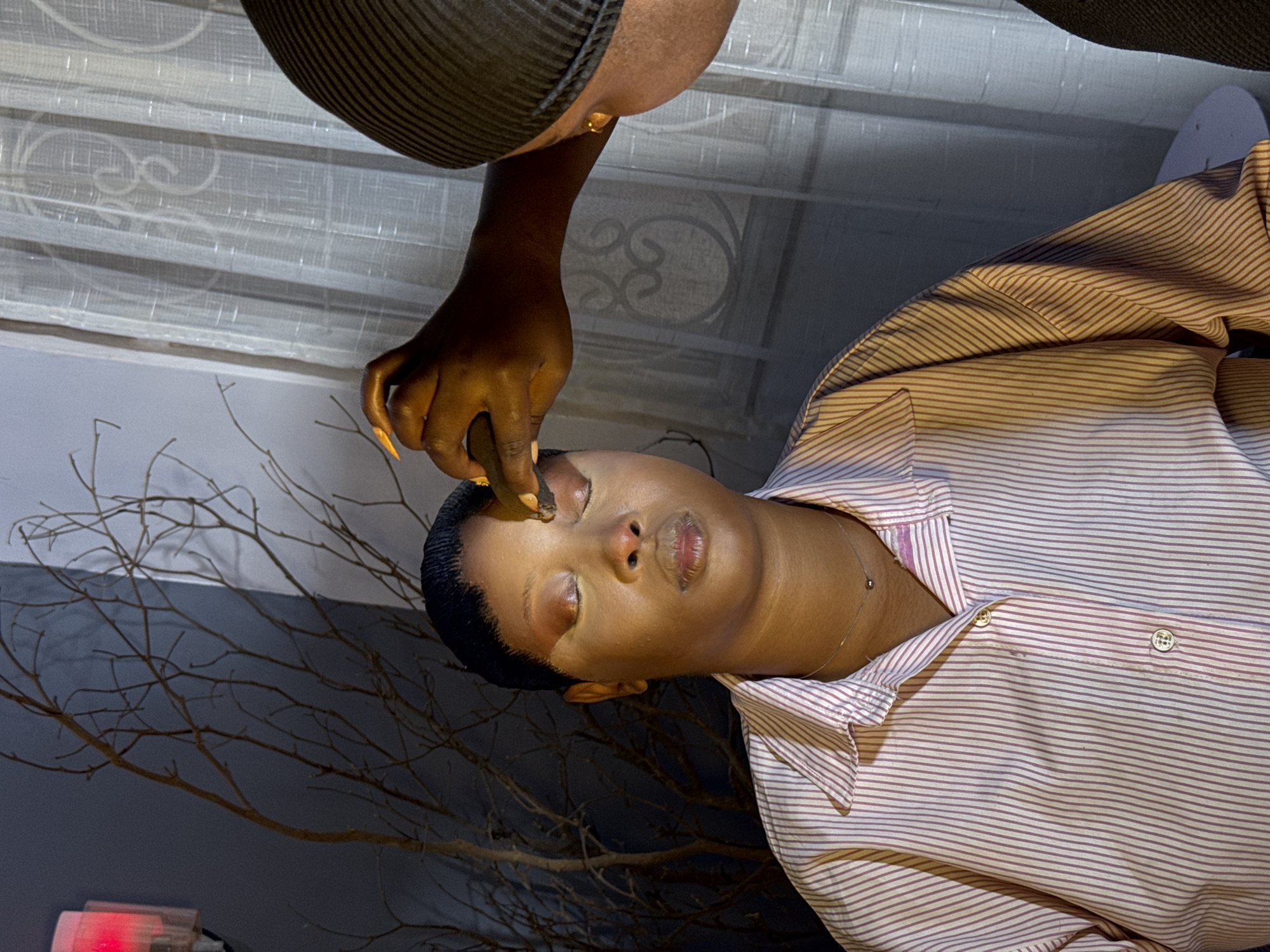 The media file shows a close-up of two individuals. One person, with dark skin and short dark hair pulled back, is the central focus. Their eyes are closed, and their face is angled upwards. A hand with long, light-colored fingernails is applying makeup, specifically to the eyelid area, using a dark-colored applicator. This individual is wearing a light-colored shirt with thin reddish-brown stripes and a delicate silver necklace.
The second individual is only partially visible, with their hand and arm in the frame, also with dark skin. They are wearing a dark, ribbed hat and a gold earring. The background consists of sheer, white curtains with decorative patterns, suggesting an indoor setting, possibly a home. A bare, branched tree is visible in the lower portion of the frame, adding an element of nature to the scene. A small, pinkish-red light source is also visible in the lower left corner. The lighting appears to be artificial, casting soft shadows and highlighting the makeup application. The overall mood is intimate and focused, likely capturing a moment of personal grooming or preparation. There is no visible text in the media file.