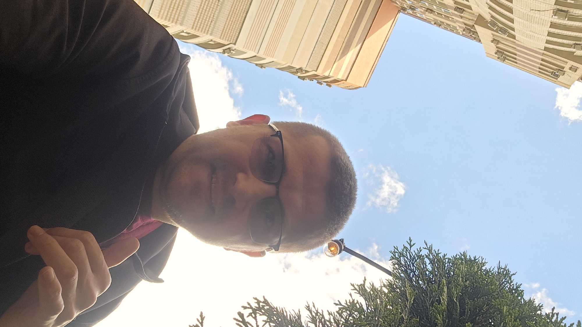 A low-angle, self-portrait captures a man with short hair and glasses, wearing a dark jacket over a pink shirt, looking up towards the sky. His right thumb is raised in a "thumbs up" gesture. To the left, a modern, multi-story building with a beige and orange facade dominates the frame, its windows visible as dark rectangular shapes. The background is a clear blue sky with scattered white clouds. To the right, the green branches of a coniferous tree extend into the frame, partially obscuring a string of small, orange lights hanging from an unseen fixture. The overall scene suggests an outdoor urban setting, likely in Kyiv, Ukraine, on a bright day.