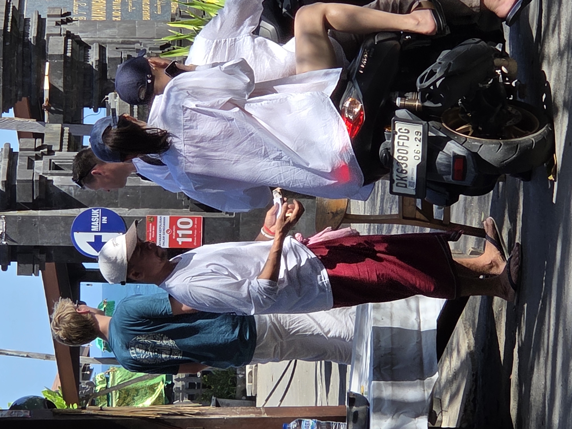 A group of people are standing and sitting around a street in North Kuta, Indonesia. In the foreground, a man wearing a white long-sleeved shirt and a maroon sarong stands and appears to be handing something to another person. Behind him, a person on a black motorcycle is seated, wearing a white shirt and a blue baseball cap. To the left, a young man in a teal t-shirt and white pants stands facing away from the camera. In the background, a street sign with an arrow and the word "MASUK IN" is visible, along with architectural structures and foliage. The scene is illuminated by bright sunlight.