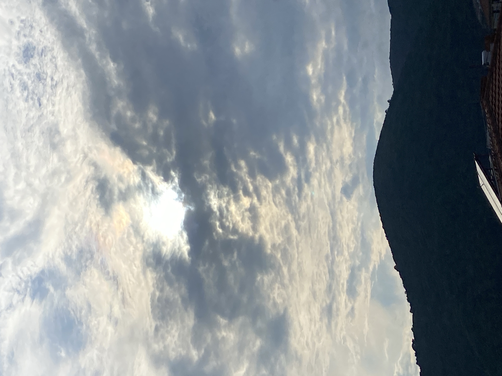 The image is a vertical, low-angle shot of a cloudy sky over a dark, silhouetted mountain range in Budva, Montenegro. The sun is partially visible through the clouds, casting a bright light and creating a rainbow-like effect. The clouds are varied in texture and density, with some appearing fluffy and white, while others are darker and more ominous. The mountain range dominates the lower half of the frame, its rugged outline sharply defined against the bright sky. In the foreground, the edge of a tiled roof and what appears to be a solar panel are visible, suggesting a residential area. The scene evokes a sense of dramatic weather and natural beauty.