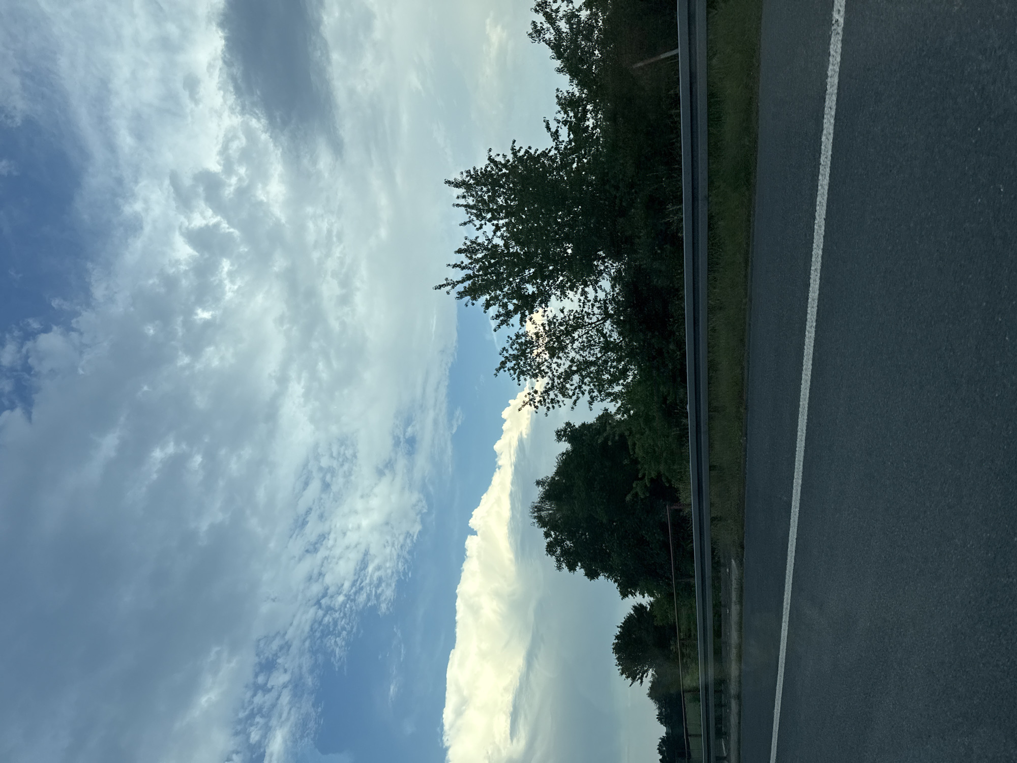 The image depicts a roadside scene in Linden, Germany. The perspective is from a vehicle, likely moving, as evidenced by the blurred motion of the road markings and the perspective of the trees along the side. The foreground is dominated by a dark asphalt road with a white line marking the lane. To the left of the road, there's a metal guardrail and a grassy verge, followed by a dense line of trees. Beyond the trees, the sky is visible, filled with a dramatic display of clouds. A large, bright white cloud formation, possibly a cumulus congestus or cumulonimbus, stands out against the blue sky, suggesting a dynamic weather system. The time of day appears to be either late morning or afternoon, given the brightness of the sun, though it is partially obscured by the clouds. The overall mood is one of travel and nature, with a sense of the vastness of the sky contrasting with the man-made road and the natural treeline. There is no visible text in the image.