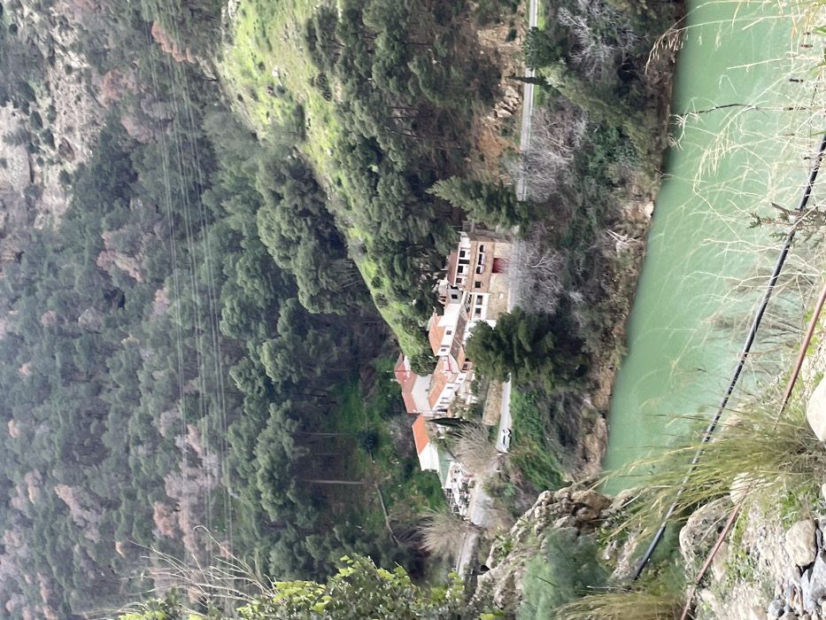 This media file captures a picturesque, albeit rotated, view of a river valley in El Chorro, Spain. The scene features a vivid, turquoise-green river flowing through the bottom right portion of the frame, flanked by steep, heavily forested hillsides. The water appears calm, reflecting the overcast or diffused daylight.
A small collection of white and earth-toned buildings with red-tiled roofs is nestled into the left bank of the river, indicating a small village or hamlet built into the mountainous terrain. A narrow road or path winds alongside these structures, leading along the river. The hillsides rise dramatically above and around these buildings, densely covered with a mix of evergreen trees and some areas showing sparser vegetation or rocky outcrops.
In the upper left quadrant, several parallel lines, likely power lines or cables, stretch across the valley, suggesting utility infrastructure in the area. The lighting is soft and diffuse, indicating an overcast day without strong shadows. No people or specific activities are discernible, and there is no visible text in the image. The overall impression is one of a tranquil yet rugged natural landscape with human dwellings integrated into its dramatic contours.