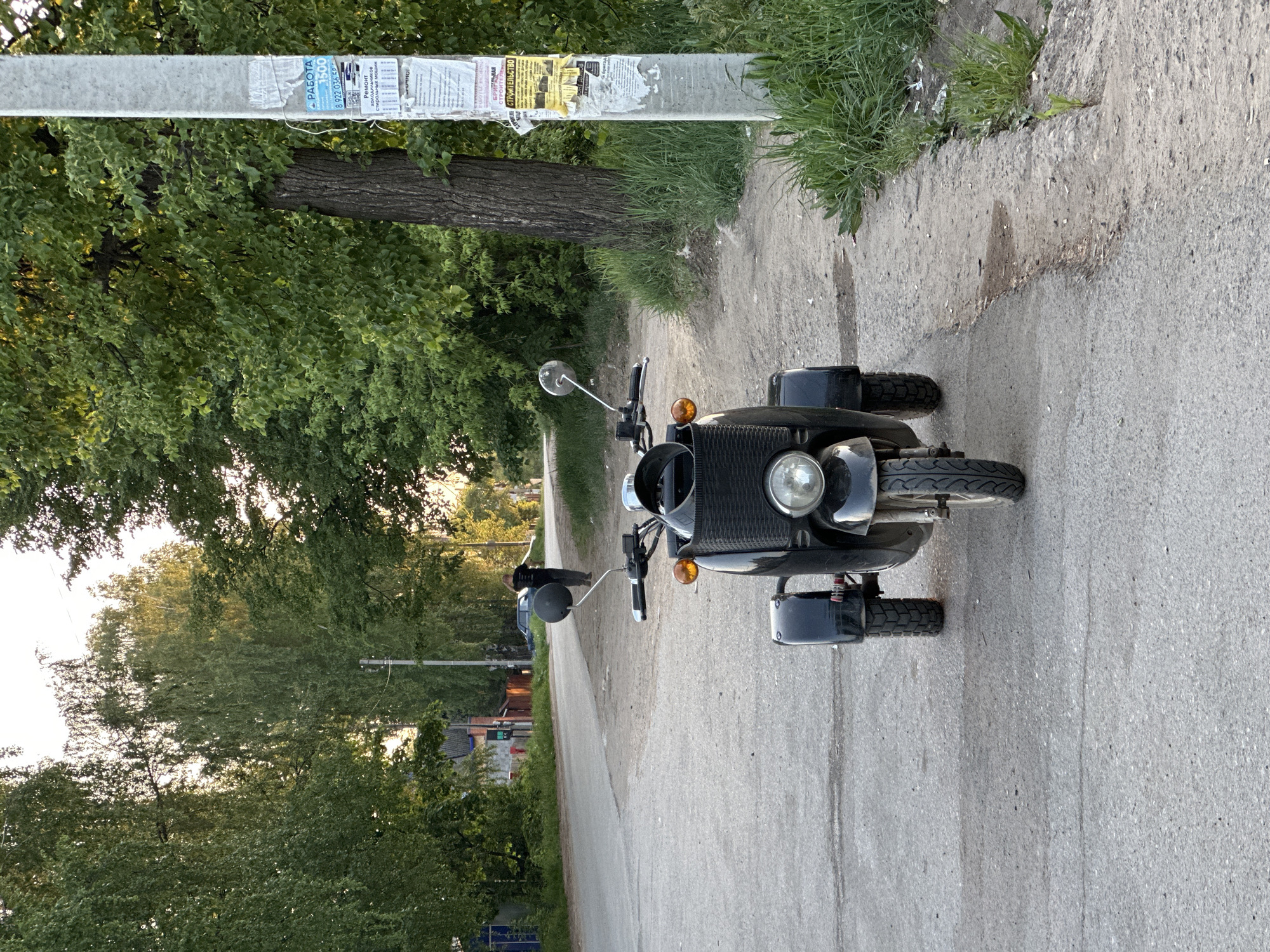 A black motorcycle with a sidecar is parked on a narrow asphalt road in what appears to be a residential area of Chaikovsky, Russia. The motorcycle is facing the viewer, with its headlight and front wheel prominently visible. The sidecar is on the left side of the motorcycle. To the left of the road is a line of trees and vegetation, suggesting a park or green space. A utility pole with posters attached to it stands next to the road on the left side of the image. A person is walking away from the motorcycle, farther down the road in the distance. The road is flanked by sidewalks or grassy areas. The overall setting conveys a calm, possibly suburban atmosphere in the city of Chaikovsky.