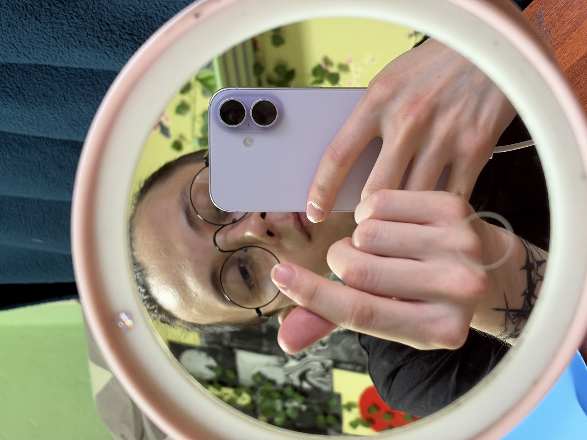 This is a reflection in a ring light of a person taking a selfie. The person is wearing round, wire-rimmed glasses and has short, dark hair. They are holding a light purple smartphone horizontally with their right hand, positioning it to capture the reflection. Their left hand is visible, with fingers pointing towards the phone. A black tattoo with sharp, angular designs is visible on their left wrist. The ring light itself has a pale pink rim.
In the background, reflected in the mirror, is a wall with yellow wallpaper featuring a floral pattern. There are also some green plants and what appear to be framed pictures or posters on the wall. The overall scene suggests an indoor setting, possibly a room in a home. The lighting appears to be artificial, consistent with the use of a ring light. The image is oriented vertically, indicating the original photograph was taken in portrait mode. There is no discernible text in the image.