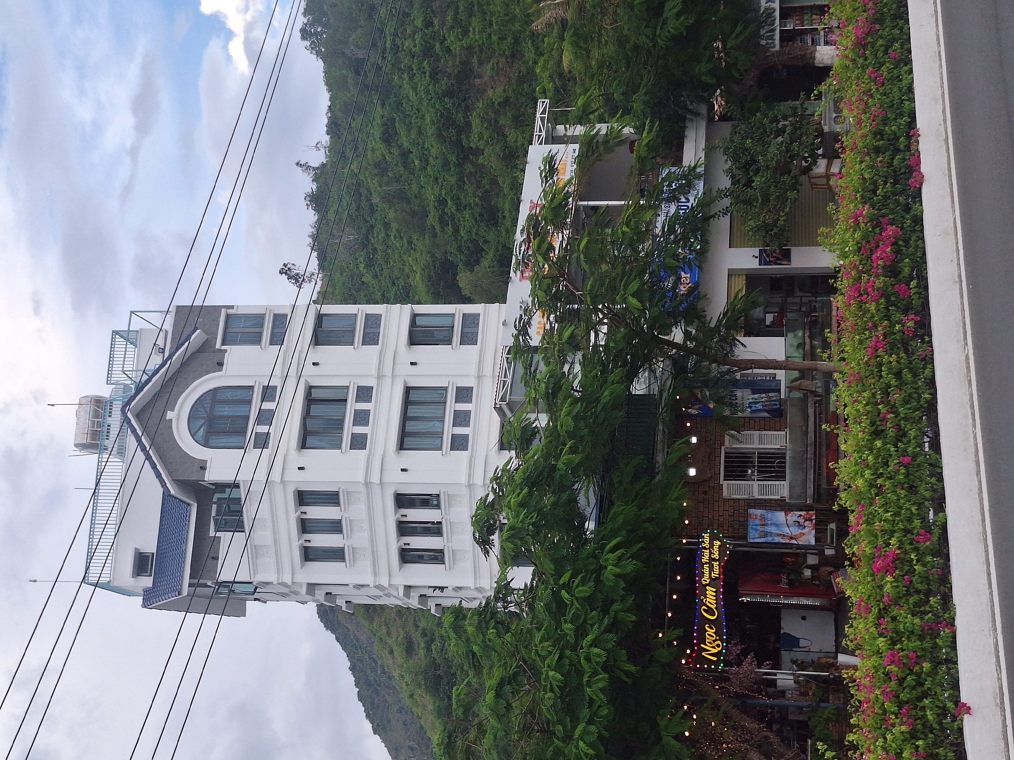 The image depicts a multi-story white building, possibly a hotel or apartment complex, situated on a hillside in Nha Trang, Vietnam. The building is modern in design with a blue tiled roof and several windows. In front of the building, there is a row of trees and a vibrant flower bed. A neon sign with Vietnamese text, "Ngọc Cẩm Quán Hải Sản Tươi Sống," is visible on a brick structure in the foreground, indicating a seafood restaurant. The sky is overcast, and power lines are prominent in the upper portion of the image. The scene suggests a commercial area alongside a residential or hospitality establishment, with lush green foliage characteristic of a coastal region.