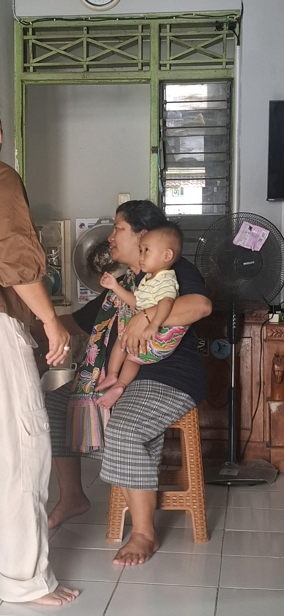 A medium shot captures an indoor scene in Desa Gagaksipat, Indonesia, where an older woman is holding a young child. The woman, seated on a brown woven plastic stool, wears a black t-shirt and plaid pants. She cradles a baby dressed in a striped shirt and colorful patterned shorts. The baby's feet are bare and rest on the woman's lap. Beside the woman, a second person, partially visible on the left, wears a brown top and light-colored pants, with their hands in view. The background features a green-painted window frame with decorative cross-hatching, a metallic fan on the right, and wooden furniture. The floor is tiled with light gray squares.