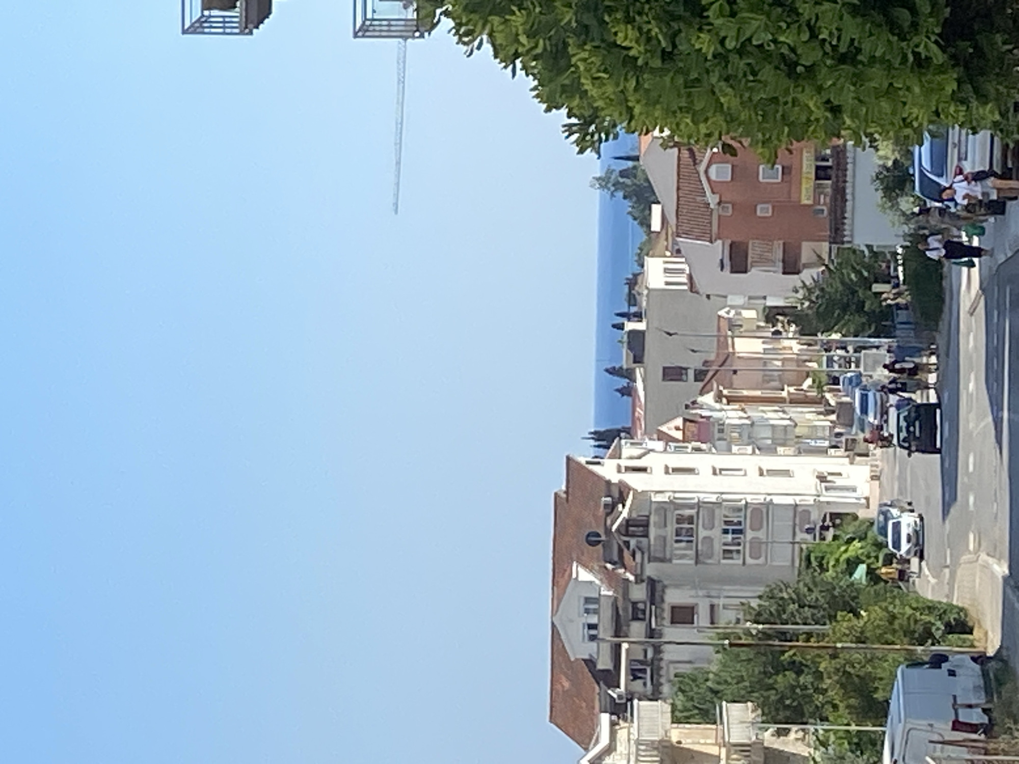 The image depicts a street scene in Budva, Montenegro, on a clear, sunny day. The perspective is from a slightly elevated position looking down a street lined with multi-story buildings. The buildings are a mix of architectural styles, with some featuring terracotta roofs and others modern facades with balconies.
In the foreground, lush green trees line the street, partially obscuring the view of the buildings. A street with parked cars and some moving vehicles is visible. Several people are seen walking along the sidewalk and crossing the street. One group appears to be near a white van, possibly interacting with it. The style of dress suggests casual summer attire.
Further down the street, the buildings continue, leading towards a distant view of the sea. The water is a calm, deep blue, and the horizon is visible under a clear sky. In the far background, a tall construction crane is visible, indicating ongoing development or a large construction project in the vicinity.
The overall impression is a lively, urban setting on a warm, bright day, with a juxtaposition of residential and commercial buildings against the backdrop of the Adriatic Sea. There is no visible text in the image.