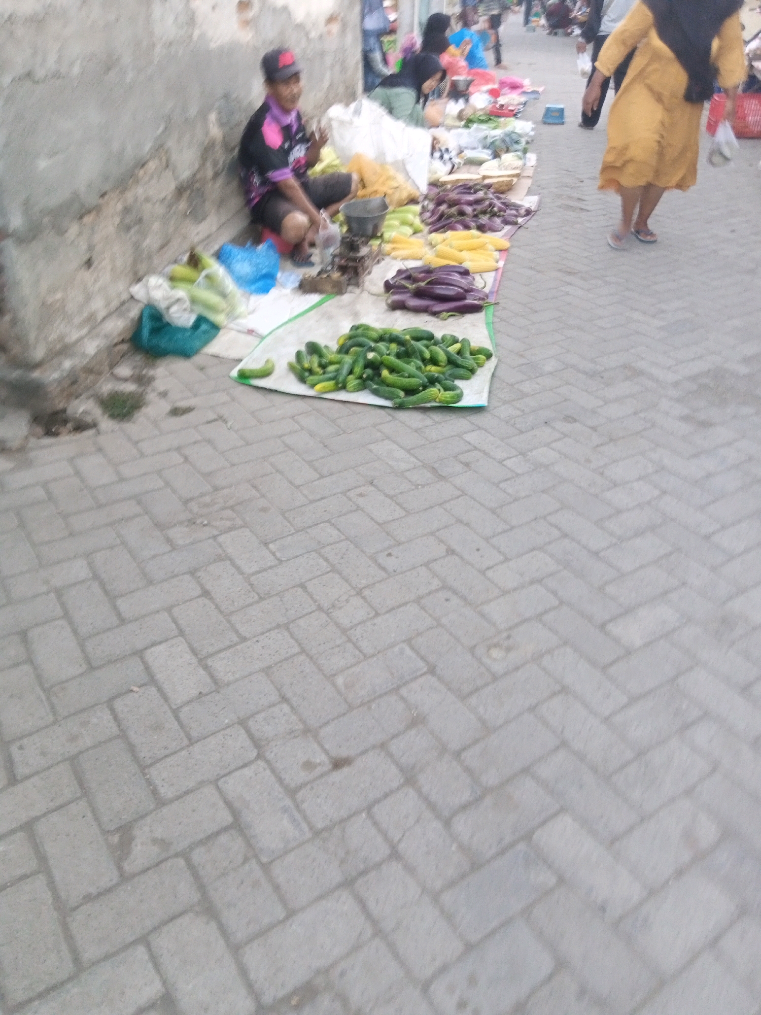 In Sono Ageng, Indonesia, at a street market, a vendor wearing a cap and a pink and black shirt sits by a wall, gesturing with their right hand while looking at the camera. In front of the vendor are vegetables, including corn, eggplants, and cucumbers, laid out on white fabric. A scale and a metal bowl are placed on the ground next to the vendor. Further down the market, people are seen browsing the stalls. A woman in a yellow dress walks away from the camera, carrying a black bag on her back. The market is set up on a paved area with a herringbone brick pattern.