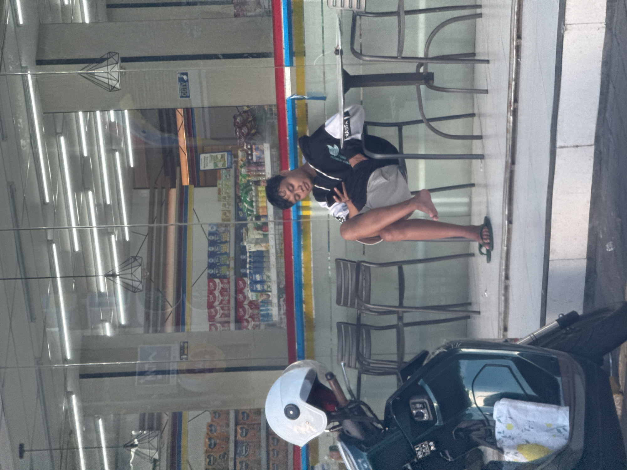 A young man is captured posing in front of a glass-fronted convenience store or minimart, likely in Kuta, Indonesia. The image, originally rotated ninety degrees clockwise, shows the man seated on an outdoor metal chair, smiling and making a peace sign with his right hand. He is wearing a black and white hooded jacket with white text "XAEJ" (partially visible) on the sleeve, light-colored shorts, and one black flip-flop on his right foot, while his left foot is bare. His posture is relaxed, with one leg bent and the other extended.
In the foreground, a dark-colored scooter with a white helmet resting on its handlebars/seat is parked. A white bag with yellow motifs is placed on the scooter's seat. Through the large glass windows of the store, the interior is visible, featuring long fluorescent tube lights on the ceiling, modern geometric pendant light fixtures, and shelves stocked with various retail products. Several metal chairs and tables, similar to the one the man is using, are also visible inside the store through the glass. A blue sign with "CCTV" and a camera icon is faintly visible on an interior wall. Vertical red, blue, and yellow stripes adorn part of the building's facade, either as decoration or structural elements. The setting appears to be daytime, with a generally bright but indirect light source. The overall atmosphere suggests a casual urban or suburban environment.