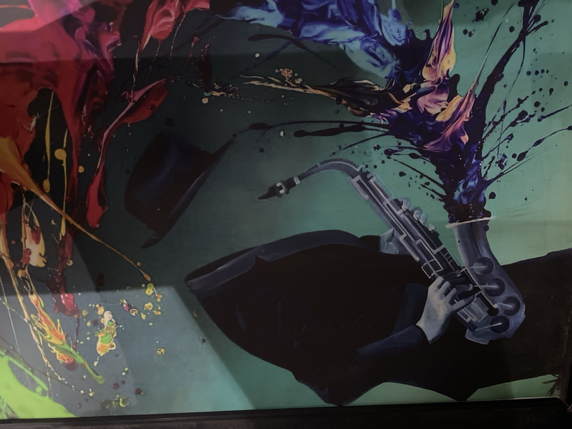 The image depicts an artistic painting of a figure playing a saxophone, set against a vibrant, abstract background. The figure is dressed in a dark suit and hat, with their face obscured by shadows. The saxophone is detailed, with a silver finish and dark keys. The background features a splash of colorful paint, with hues of green, yellow, red, pink, and purple, creating a dynamic and energetic feel. There are also small splatters of paint scattered across the canvas. The painting is framed in black. The image does not contain any identifiable landmarks or elements that would suggest a specific location within the city of Abuja, Nigeria.