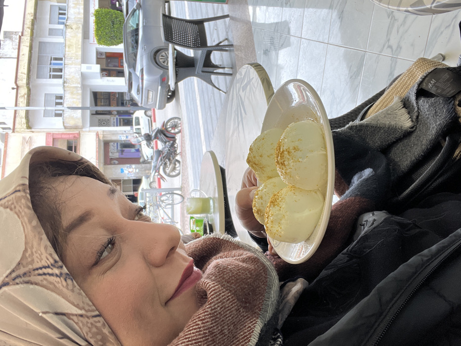 The image depicts an indoor scene looking out onto a city street in Meknes, Morocco. In the foreground, positioned to the left, an adult female is partially visible from the chest up, looking downwards with a slight smile. She is wearing a beige headscarf with a brown pattern, a thick brown and black striped scarf, and a dark outer jacket. Her right hand is holding a white oval plate containing four uniform, off-white, semi-spherical food items, each dusted with a golden-brown powder on top. To her immediate right, a light-colored round table holds a transparent green glass mug filled with a pale yellow-green liquid.
Through a large glass window, a street scene unfolds under daylight. The background features two-story commercial buildings with light-colored facades, some exhibiting architectural details. Parked vehicles include a dark gray SUV (Peugeot 3008 or similar), a black motorcycle, a white sedan, and a bicycle. A black outdoor table with a matching black wicker-style chair is visible on the tiled pavement outside. The street is paved, and a street light pole stands between the outdoor cafe seating and the roadway. Shopfronts with indistinct signage are visible along the street. The indoor flooring consists of light-colored tiles.