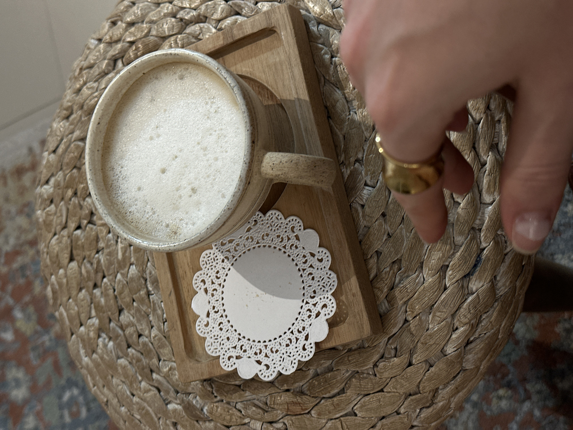 A high-angle shot displays a mug filled with a light-colored, frothy beverage set on a small wooden tray. A delicate white doily lies between the mug and the tray. The mug rests on a round, woven stool-like surface. A hand with a gold ring is partially visible in the upper right, appearing to reach towards the mug. The location is assumed to be in Kerobokan Kelod, Indonesia.