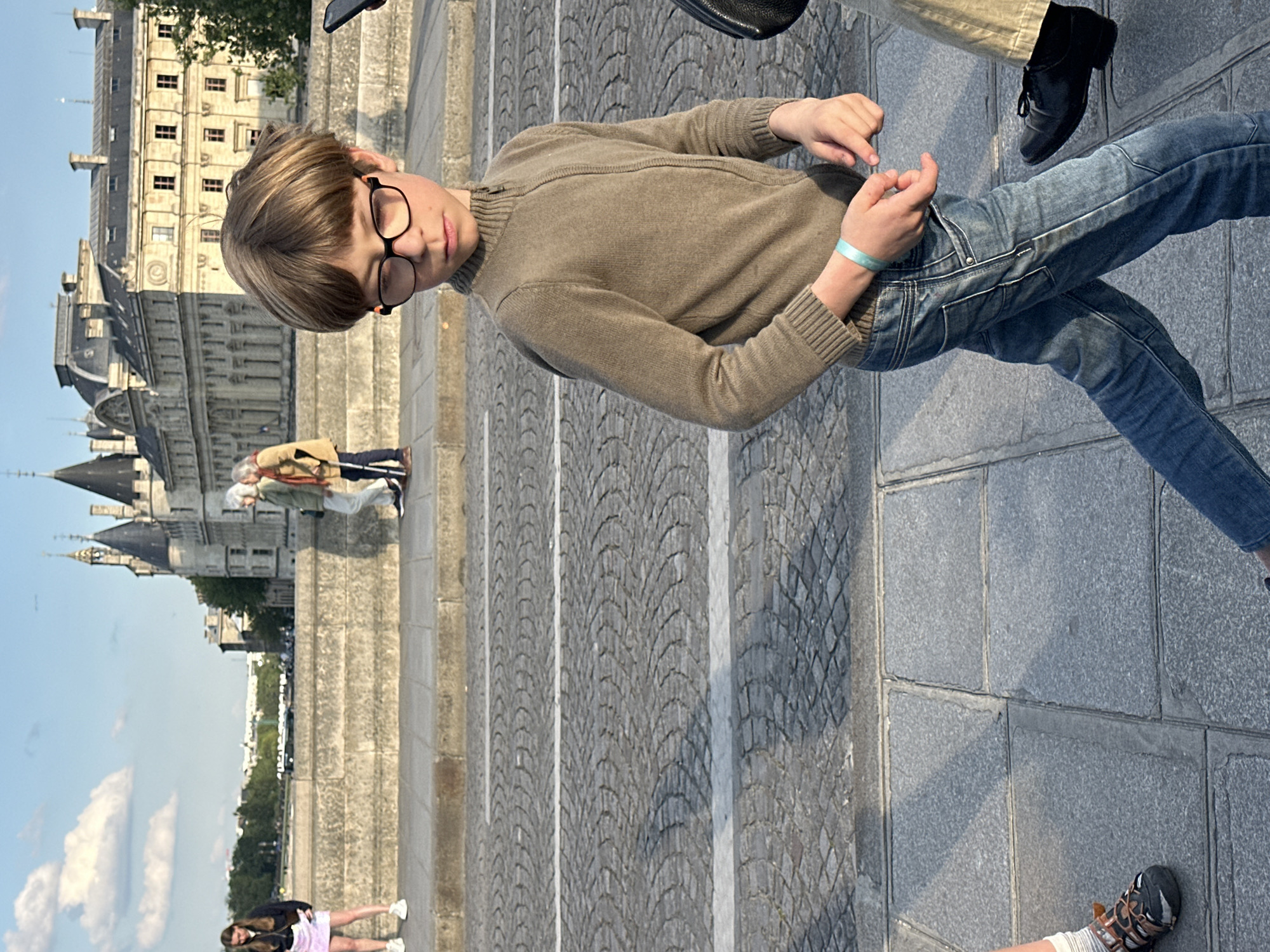 The image shows a young person wearing glasses, a brown sweater, and jeans standing on a cobblestone street in Paris. In the background, the Conciergerie, a former royal palace and prison, is visible. Other people are also in the vicinity, and the scene appears to be outdoors on a sunny day. The young person is touching their fingers together.