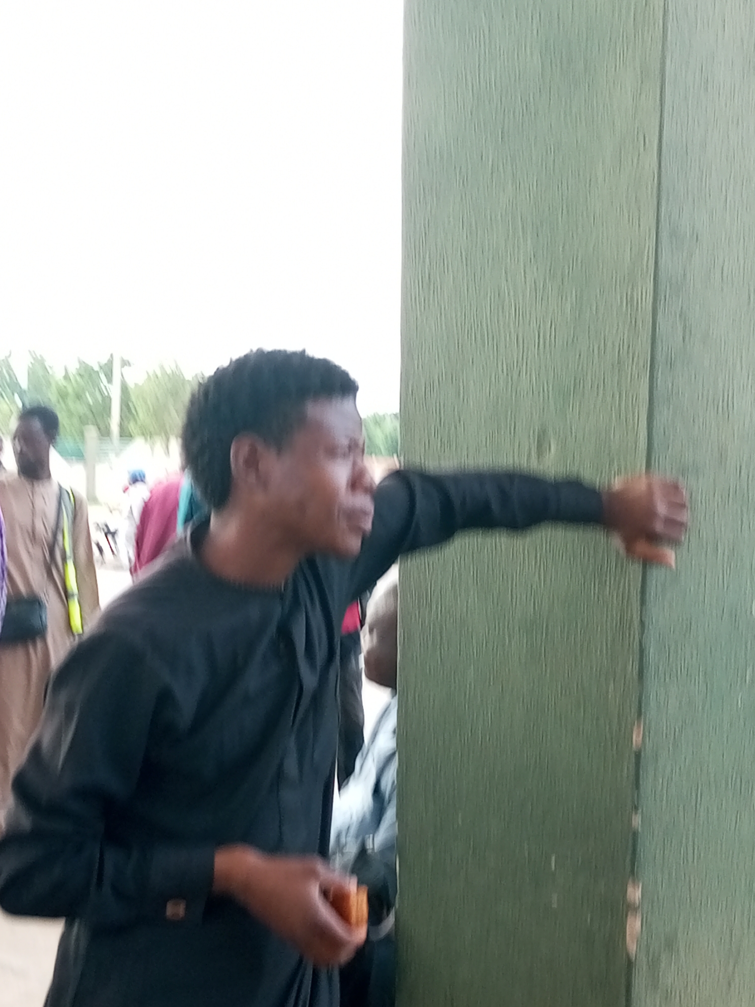 In Gombe, Nigeria, a young Black male, appearing to be in his late teens or early twenties, stands at an angle, facing right and looking away from the camera. He is wearing a black, long-sleeved garment with a mandarin collar and a cufflink visible on his left wrist. His right hand is extended and rests against a textured, pale green vertical surface, possibly a wall or door. In his left hand, he holds a small, orange, rectangular object, possibly food.
In the background, out of focus, other individuals are present. To the left, a man in a light brown garment and a yellow vest with a black crossbody bag is visible. Further in the background, other figures in colorful clothing can be discerned. The environment appears to be outdoors, with a bright, overcast sky and hints of greenery and structures in the distance. The overall scene suggests a public gathering or a casual outdoor setting.