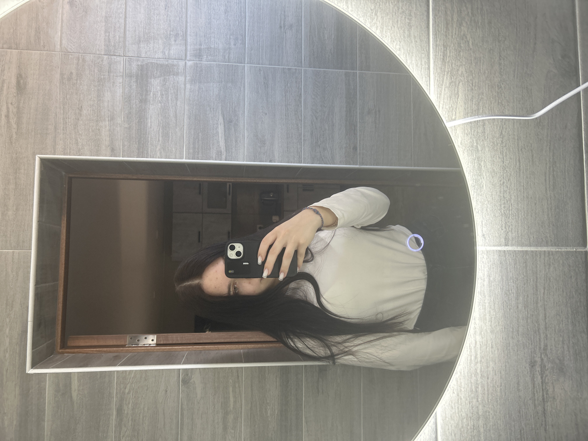 A person is taking a selfie in a bathroom mirror. The bathroom has gray, wood-look tiles on the wall. The mirror is circular with a built-in light on the right side. The person is holding a black smartphone with a white manicure visible on their nails. The reflection shows a dark-haired person wearing a white long-sleeved shirt. Inside the square-shaped part of the mirror appears to be a doorway with lockers. The location is potentially in Kemul, Russia.