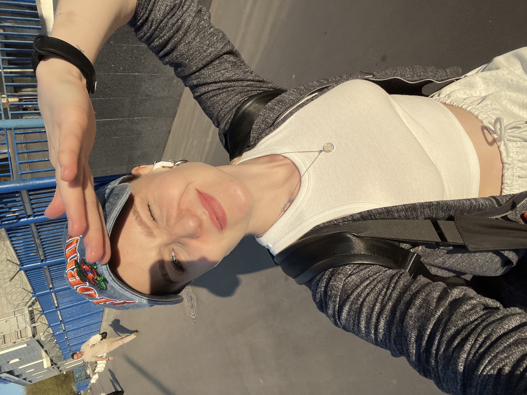A woman wearing a denim cap, a white top, and a gray zippered jacket with black straps is taking a selfie outdoors. She is shielding her face from the sun with her hand. She has a black fitness tracker on her wrist. In the background, a man in a light-colored outfit walks by. The location appears to be near blue metal railings and building structures, possibly in Khimki, Russia.