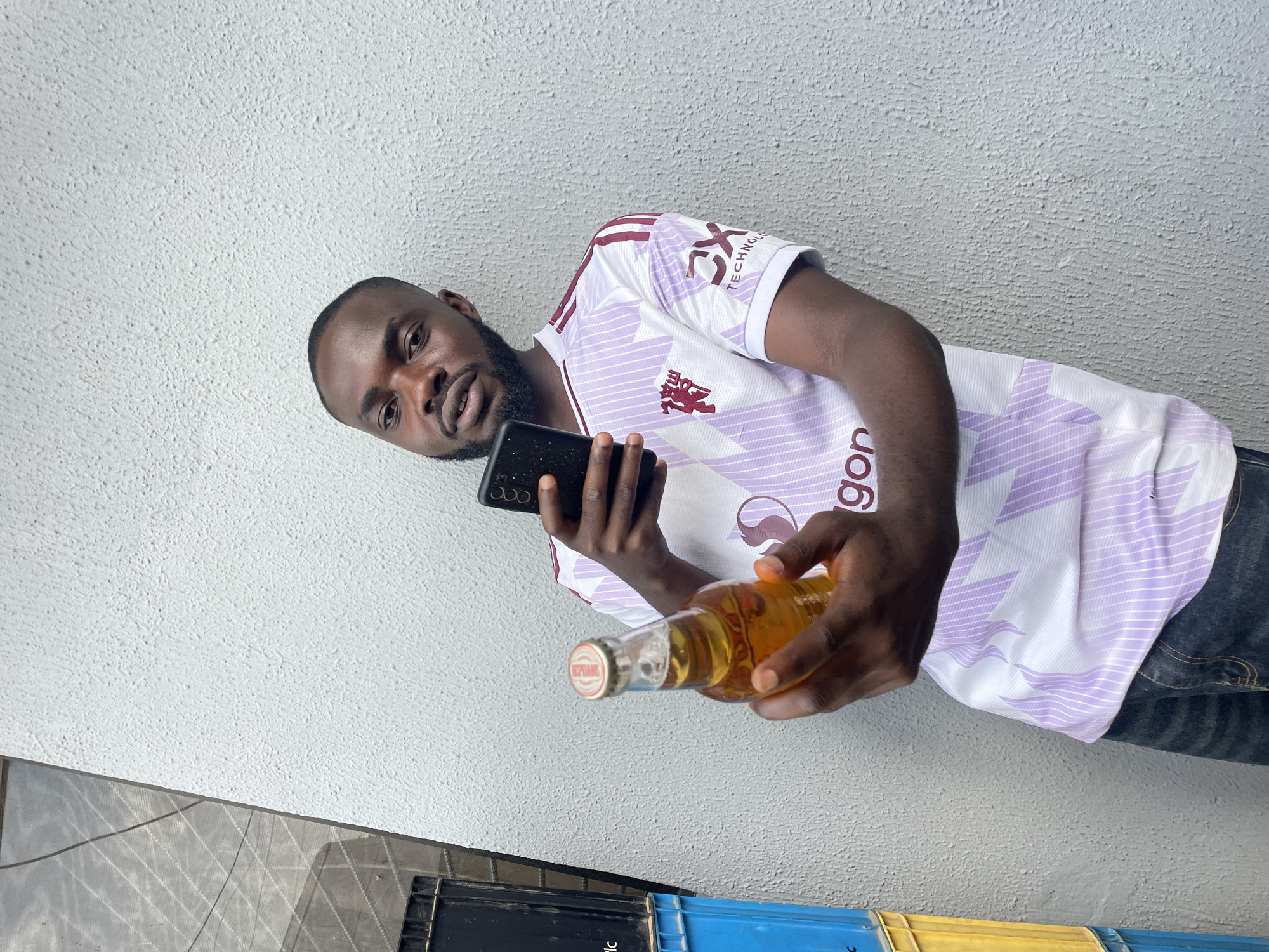 The image shows a man holding a mobile phone in his left hand and a bottle of beer in his right hand, with the bottle cap facing the camera. He is wearing a white and purple striped sports jersey with a logo on the left chest and text on the sleeve. The man has dark skin and short hair and a beard. He is looking at the camera with a neutral expression. The background is a textured white wall. The scene is outdoors, possibly in a residential area, with some plastic crates visible in the lower part of the image. The lighting suggests it is daytime. The text on the bottle cap is partially visible, showing "BEER" and a logo. The text on the jersey sleeve is "DX TECHNOLOGIES". The overall impression is casual and relaxed.