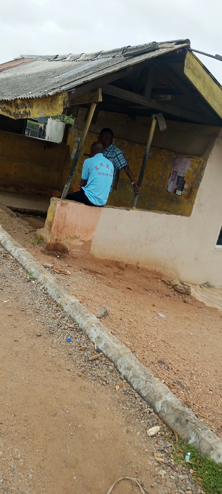 Two men are standing under a makeshift shelter with a corrugated metal roof. The shelter appears to be part of a small, rundown building painted yellow. One man, wearing a light blue t-shirt with Chinese writing and a phone number on it, is sitting on a low concrete wall. The other man, wearing a checkered shirt, stands behind him, holding onto a support pole. A poster with a portrait of a man is visible on the yellow wall behind them. The ground in front of the shelter is sandy and covered with gravel, with a concrete kerb running along the side. The scene is located in Adankolo, Nigeria.