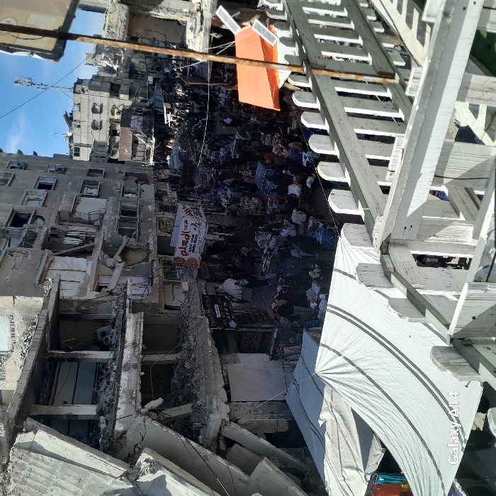 The image depicts a crowded street market amidst extensively damaged urban infrastructure in Khan Yunis, Palestinian Territory, captured from an elevated perspective. In the foreground, a white painted wooden railing runs horizontally, with a large white tarp tent, marked with two grey stripes, extending below and to the left. The bottom left corner bears a "Galaxy A14" watermark.
The midground is dominated by a bustling market scene on a paved street, densely populated by individuals navigating numerous stalls and tables laden with various goods. Many people are visible, appearing to be engaged in buying and selling activities. A prominent red and white banner displaying Arabic text, "المعلم" (Al-Mu'allim - The Teacher), hangs above the crowd, with another smaller sign, "Loyal Mobile," below it. To the right of the market, an orange tarp structure provides shelter for a stall.
The background consists of multi-story concrete buildings exhibiting severe structural damage. Multiple floors are partially or completely collapsed, exposing internal frameworks, rebar, and debris. Walls are missing, windows are shattered, and large sections of concrete are either gone or leaning precariously. Rubble is visible in various areas. The sky above is clear blue with sparse clouds. In the distance, more damaged buildings line the street, and a communications antenna is visible on a rooftop.