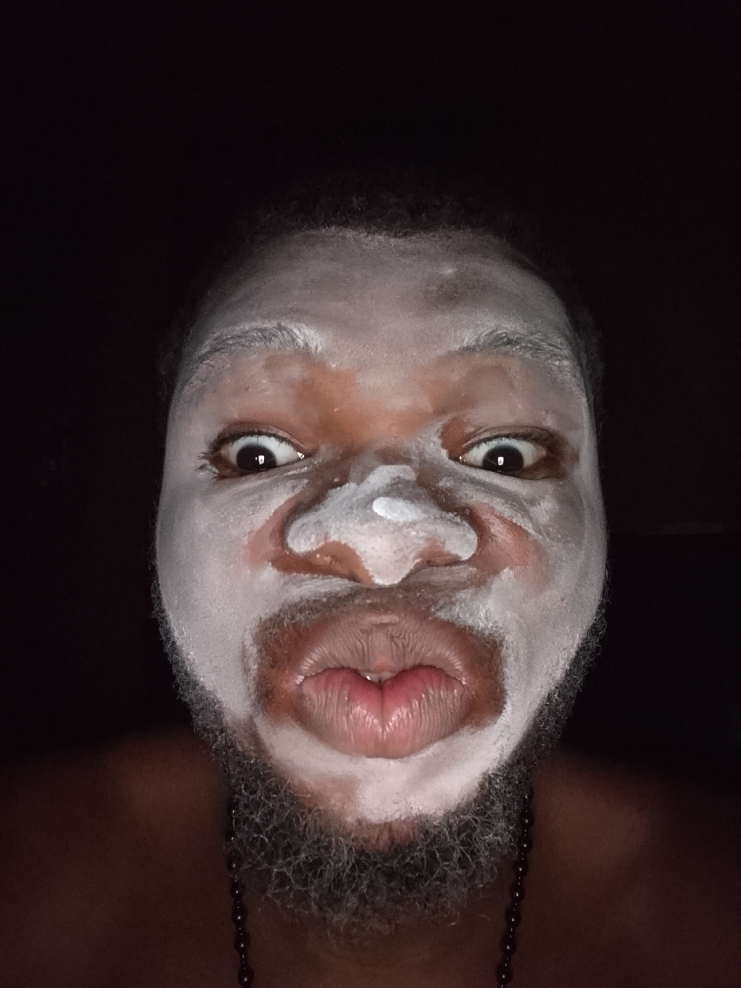 A close-up of a person's face covered in a white substance, possibly a mask, set against a dark background. The person has dark skin, a beard, and expressive eyes. They are wearing a necklace with beads. The location is unknown.