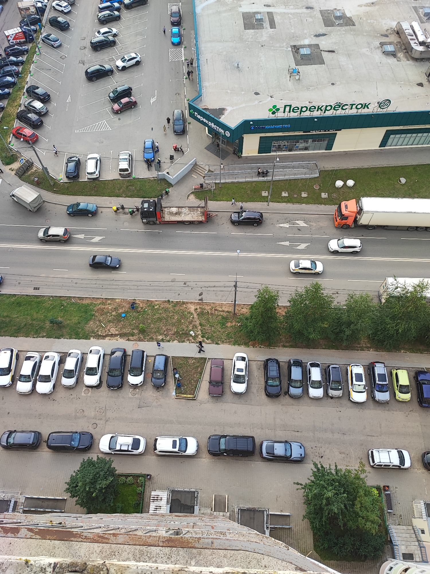 The image shows an aerial view of a parking lot and a street in Vidnoye, Russia. The parking lot is adjacent to a supermarket with a green and beige facade, identified by the "Перекрёсток" (Perekrestok) logo. Numerous cars are parked in organized rows within the lot, with a few cars also present on the street.
On the street, a flatbed truck with an orange cab is parked on the right side, facing away from the camera, with a long white trailer. Further down the street, a white semi-trailer truck is visible. Several passenger cars are driving on the street, some in motion and others parked. A few individuals are walking on the sidewalk near the supermarket entrance. The environment appears to be an urban commercial area with paved surfaces, green spaces, and a multi-story building visible in the background.