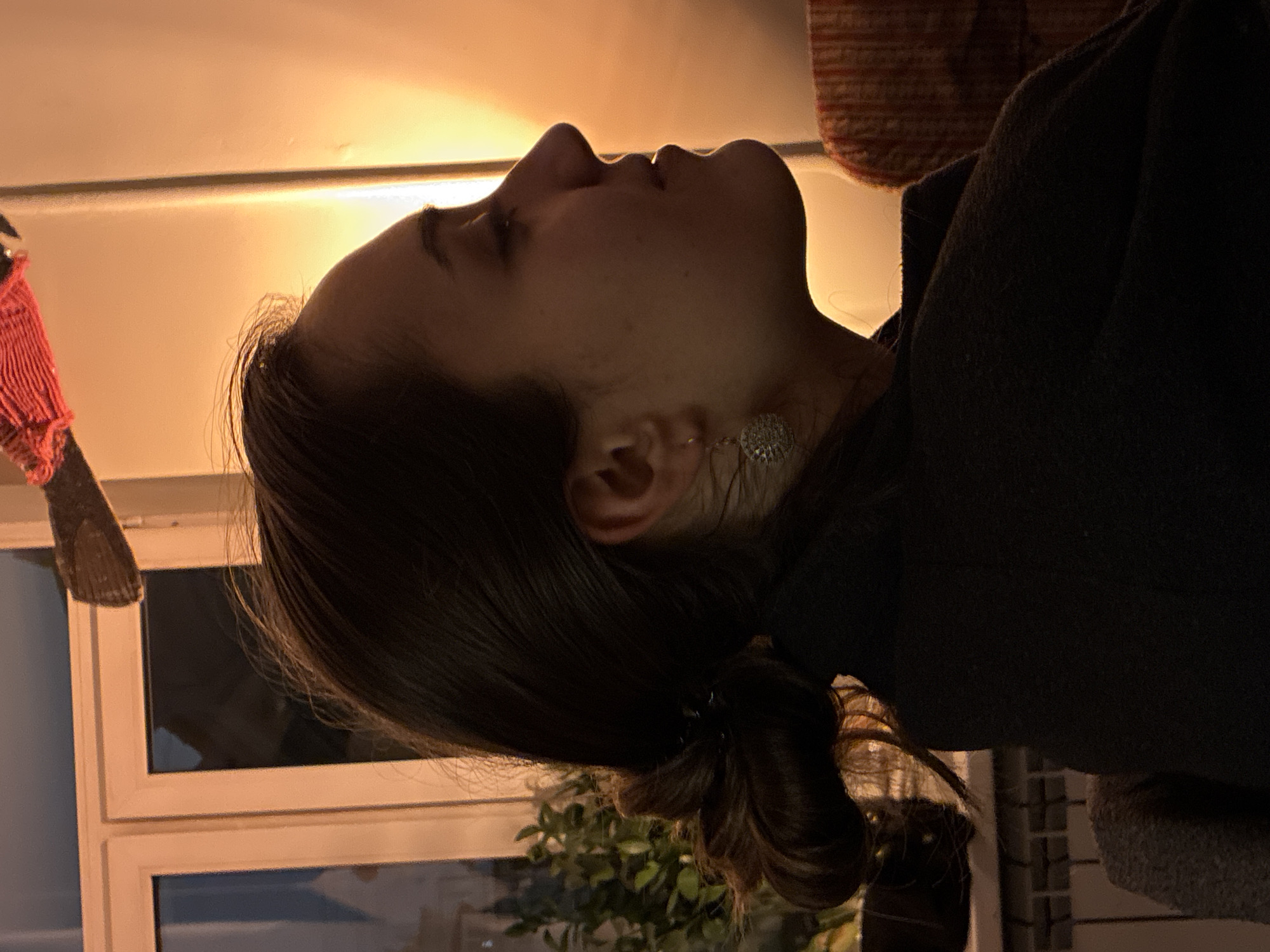 A woman with dark hair pulled back into a low bun, wearing a dark top and a dangling earring, is looking upward. She is indoors, with a window behind her showing a night sky. There is a lamp illuminating the wall behind her. A red and white object hangs to the left, and a green plant is visible behind her head. There is no information available in the image that allows a determination of location in Moscow, Russia.
