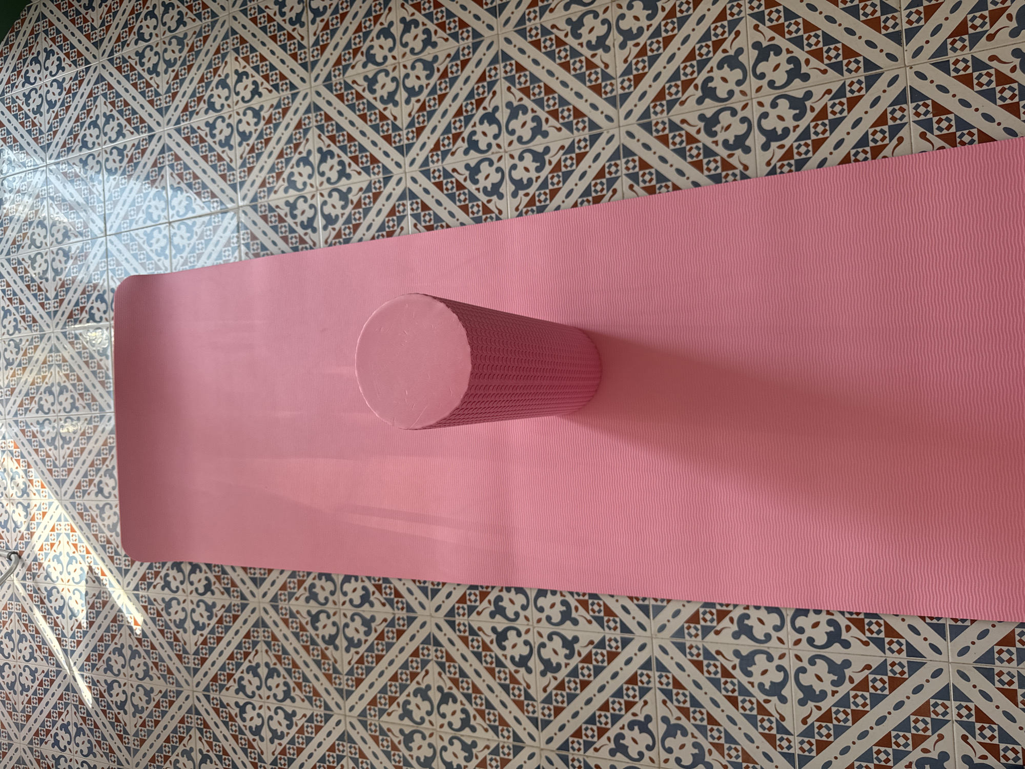 A pink yoga mat and a pink foam roller are situated on a patterned tiled floor. The tiles feature a repeating geometric design with blue, white, and brown elements. Sunlight casts diagonal shadows across the tiles and the yoga mat, indicating an indoor setting. The yoga mat is rolled out lengthwise, with the foam roller placed vertically in the middle of the mat. The image provides no information about the city of Isola delle Femmine, Italy.