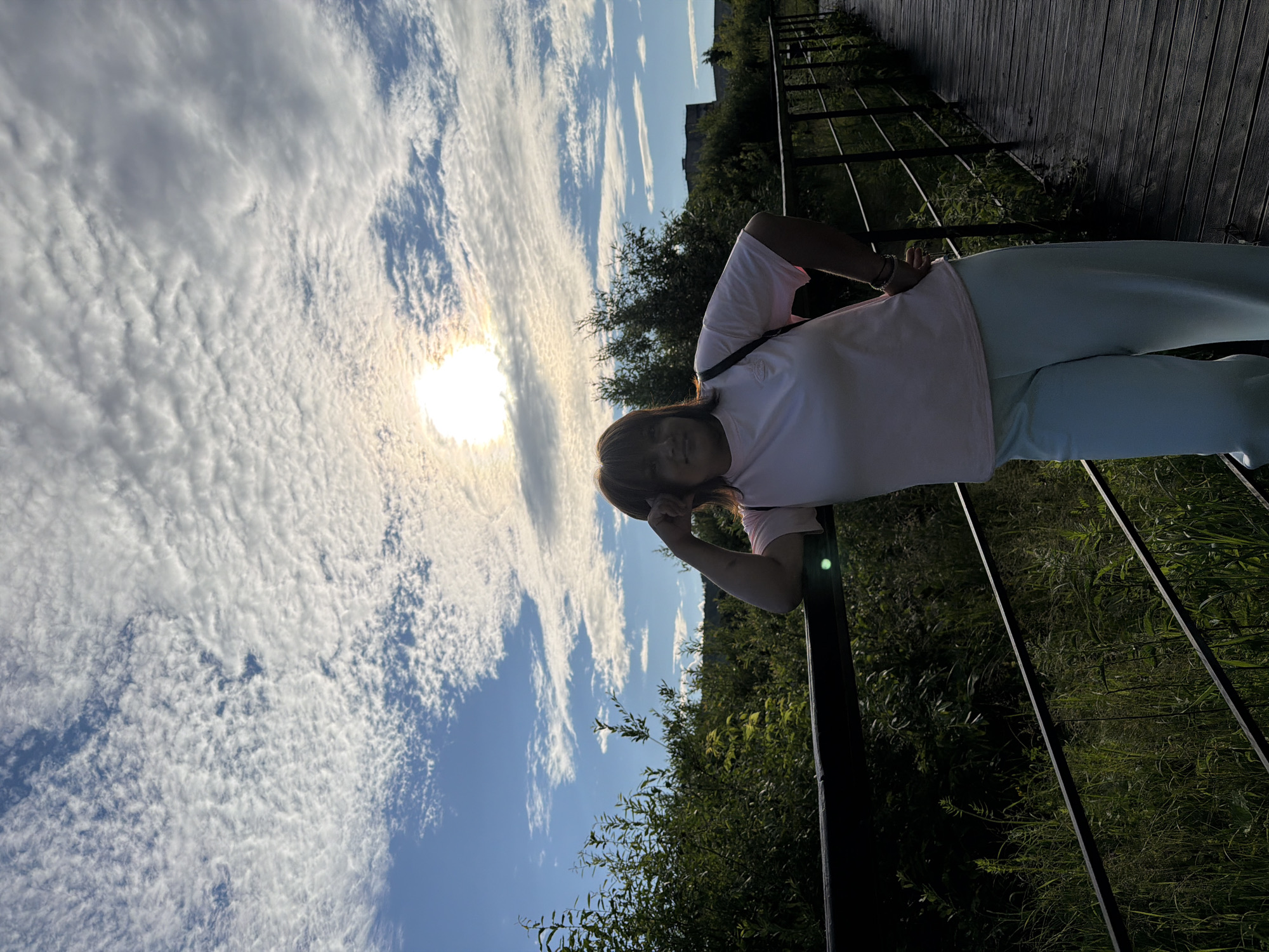 The image shows a person leaning on a railing outdoors, with a bright sun and cloudy sky in the background. The person is wearing a light pink t-shirt and light blue pants. They have dark hair and appear to be looking towards the camera, with one hand resting on their forehead and the other on the railing. The setting appears to be a boardwalk or elevated path, with wooden planks visible on the right side of the image. Lush green foliage and trees are visible below the railing, suggesting a natural environment. The sun is high in the sky, creating a bright and somewhat hazy atmosphere, indicating it is likely daytime. The sky is filled with scattered clouds, some of which are illuminated by the sun. The overall mood seems relaxed and perhaps contemplative, with the person appearing to enjoy the outdoor scenery. There is no visible text in the image.