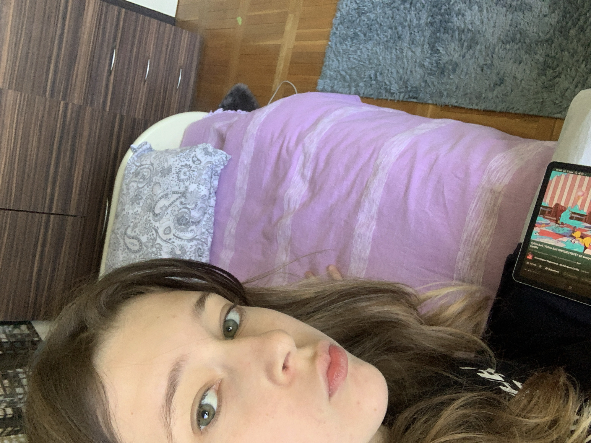 A young woman lies on a lavender bedspread with a white striped pattern. She is watching something on a tablet. A grey patterned pillow rests against the headboard. A gray rug is visible in the background. To the left is a dark brown wardrobe.
There is not sufficient information to determine if this image was taken in the city of Vozdovac Urban Municipality, Serbia.