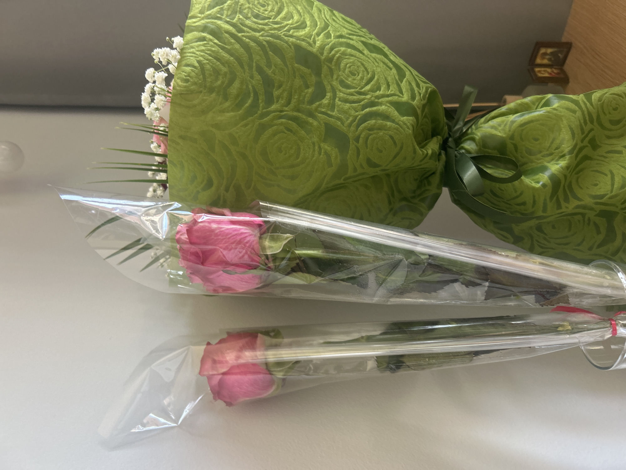 A close-up shows two pink roses individually wrapped in clear plastic sleeves, lying parallel to each other on a flat, white surface. Behind them is a larger bouquet, wrapped in green fabric with a rose pattern and tied with a green ribbon. The image is likely taken indoors, with a glimpse of a wooden wall and a framed picture in the background. There is no information indicating that this image was taken in Burgas, Bulgaria.
