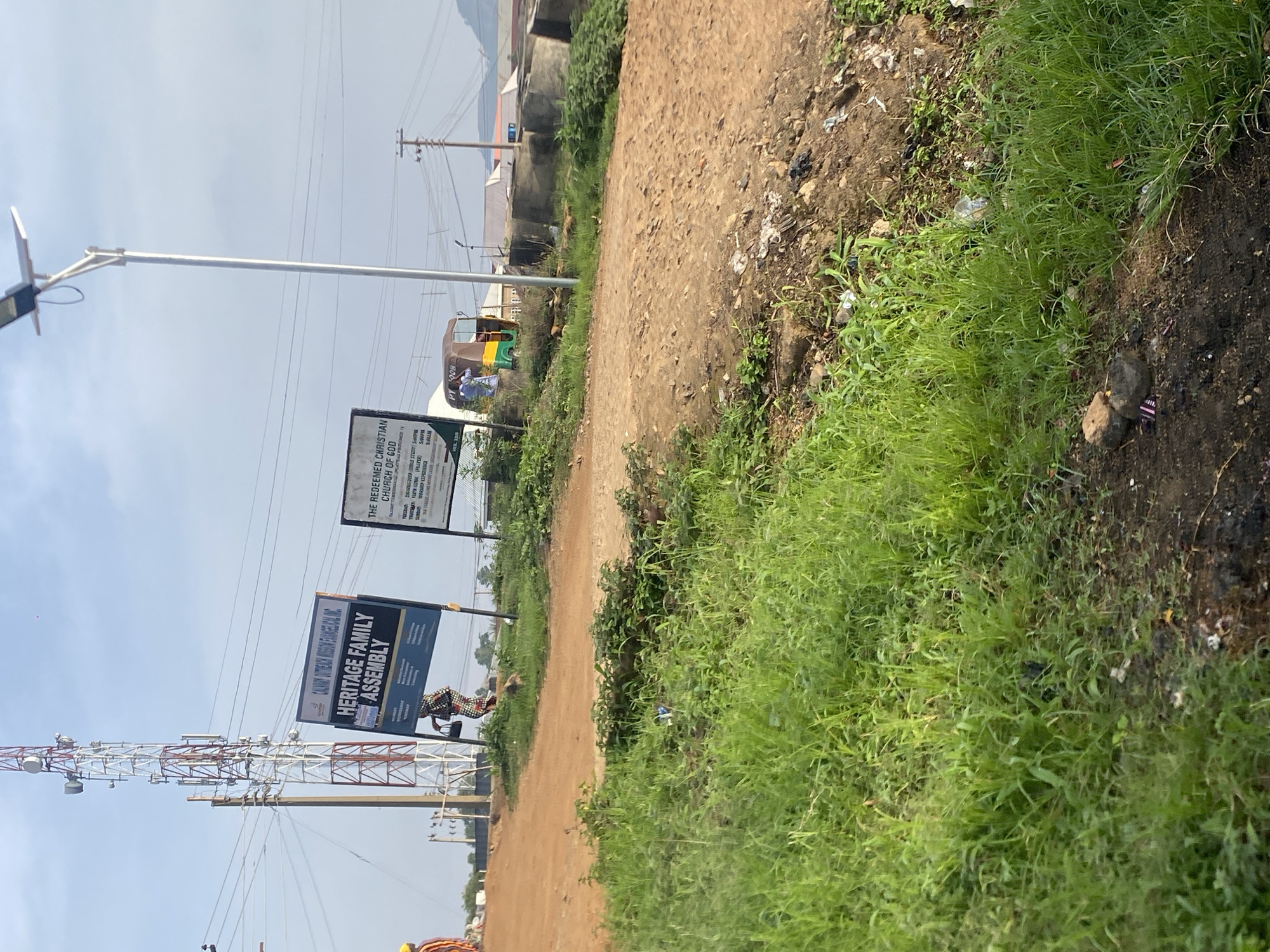 The image depicts a rural or semi-urban scene in Jos, Nigeria, characterized by a dirt road and lush green vegetation. A prominent feature is a tall communication tower, suggesting the presence of telecommunication infrastructure. Beside the tower, two signs are visible; one reads "HERITAGE FAMILY ASSEMBLY" and the other "THE REDEEMED CHRISTIAN CHURCH OF GOD". A person in traditional attire is walking towards the signs, and a three-wheeled vehicle, possibly an auto-rickshaw, is parked nearby. The environment appears somewhat unkempt, with scattered debris and unpaved surfaces. The sky is overcast, and a solar-powered street lamp stands tall, indicating ongoing development or modernization efforts. The overall impression is of a developing area with a mix of traditional and modern elements.