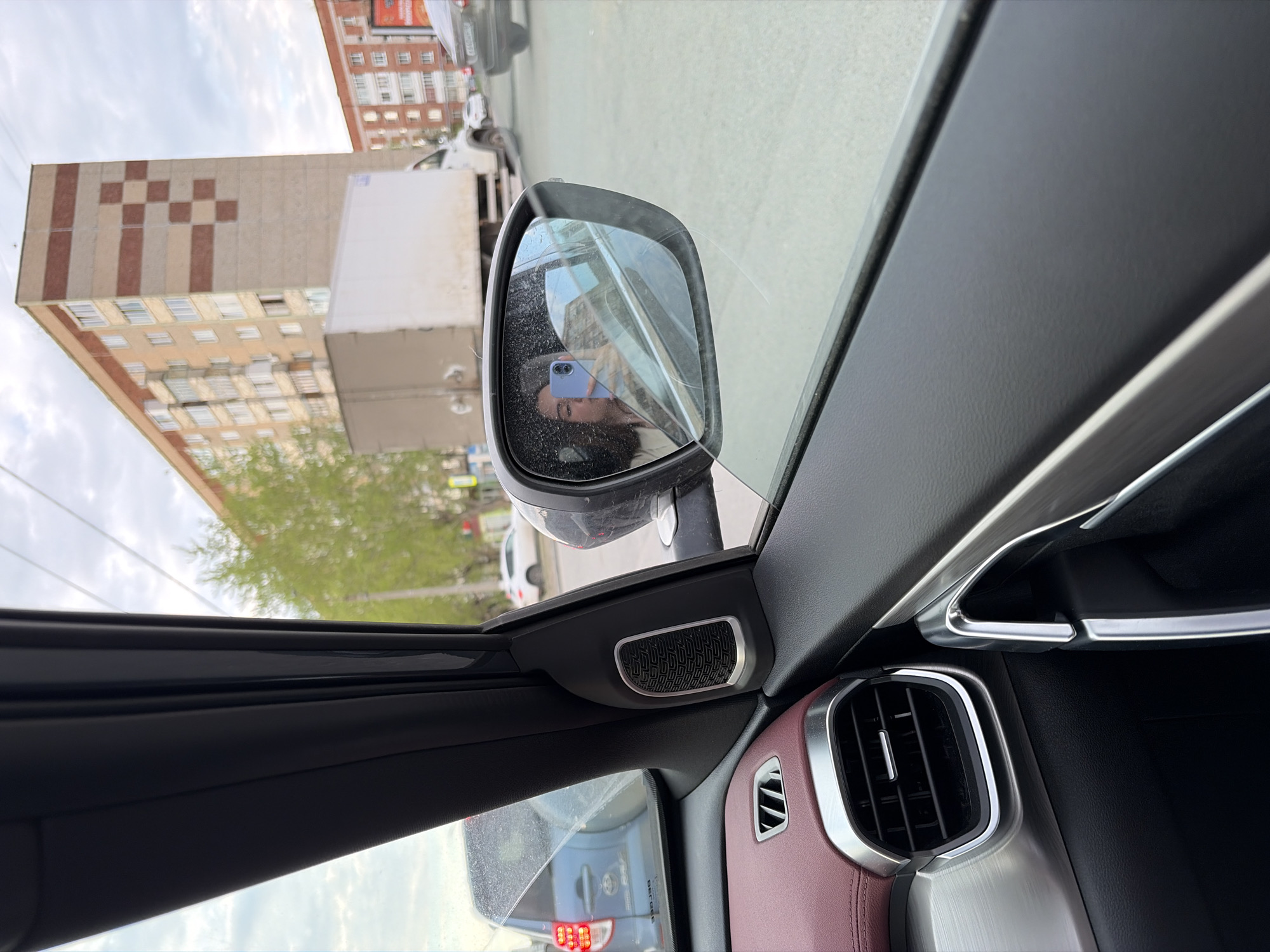 The image depicts the interior of a car, specifically a view through the driver's side window, with a focus on the side mirror reflecting the exterior environment. In the mirror, a person is visible, holding a phone. The reflection shows a building with a distinctive checkered pattern on its side, identifiable as being located in Izhevsk, Russia. Outside the car, a street scene unfolds, with buildings, vehicles, and trees. The car's interior features a red leather accent.