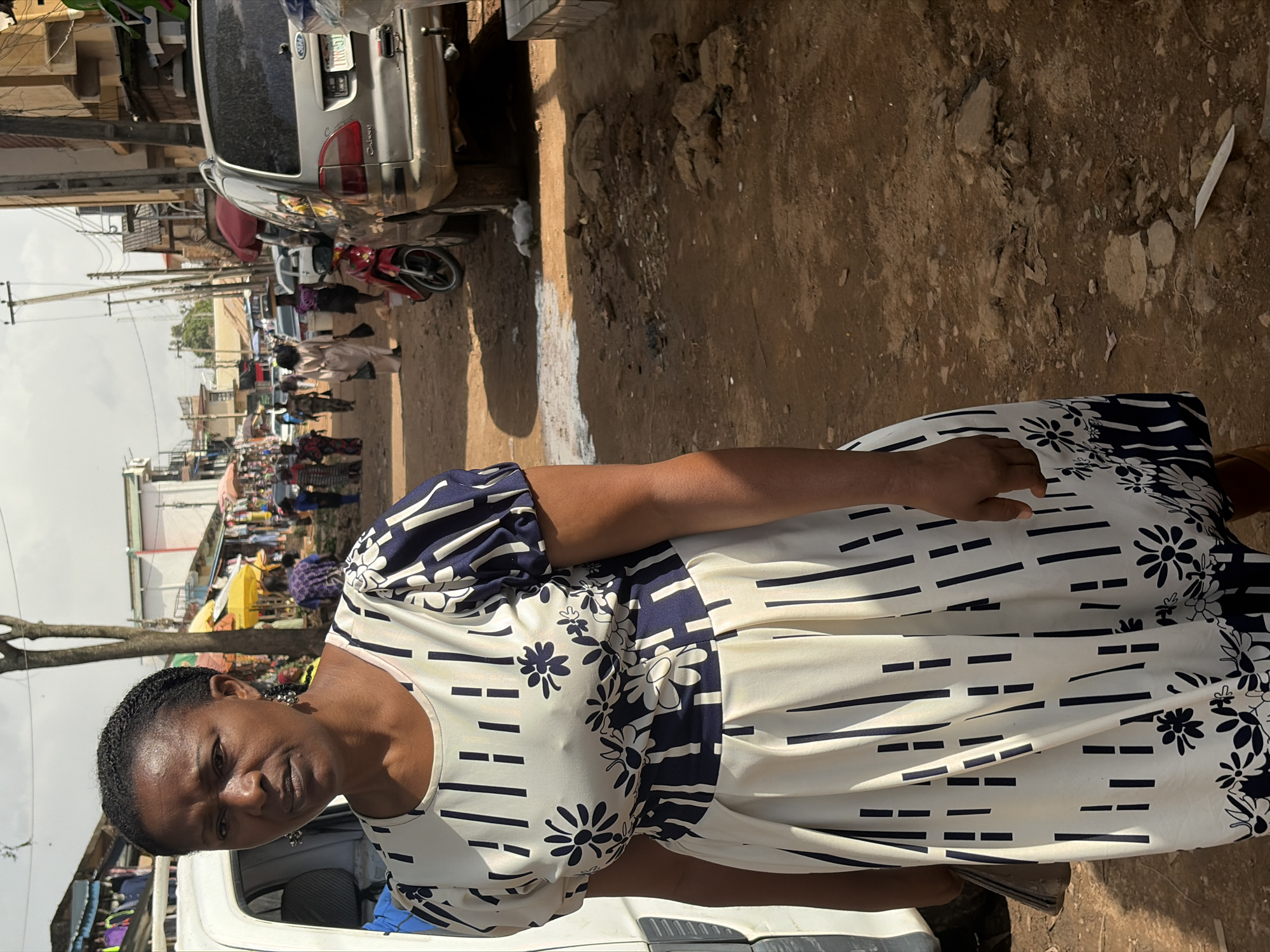 The image shows a woman in a busy outdoor marketplace. She is wearing a cream-colored dress with a navy blue pattern of stripes and flowers. The woman is looking directly at the camera with a neutral expression. In the background, there are several other people, some stalls, and vehicles, suggesting a lively commercial environment. A silver car is visible to the left of the woman. The ground appears to be dirt and gravel. The lighting indicates it is daytime, likely with bright sunlight. The setting suggests a typical market scene in Jos, Nigeria, with elements common to such locations like temporary stalls and various modes of transportation. The overall impression is of a moment captured in a bustling, everyday market.