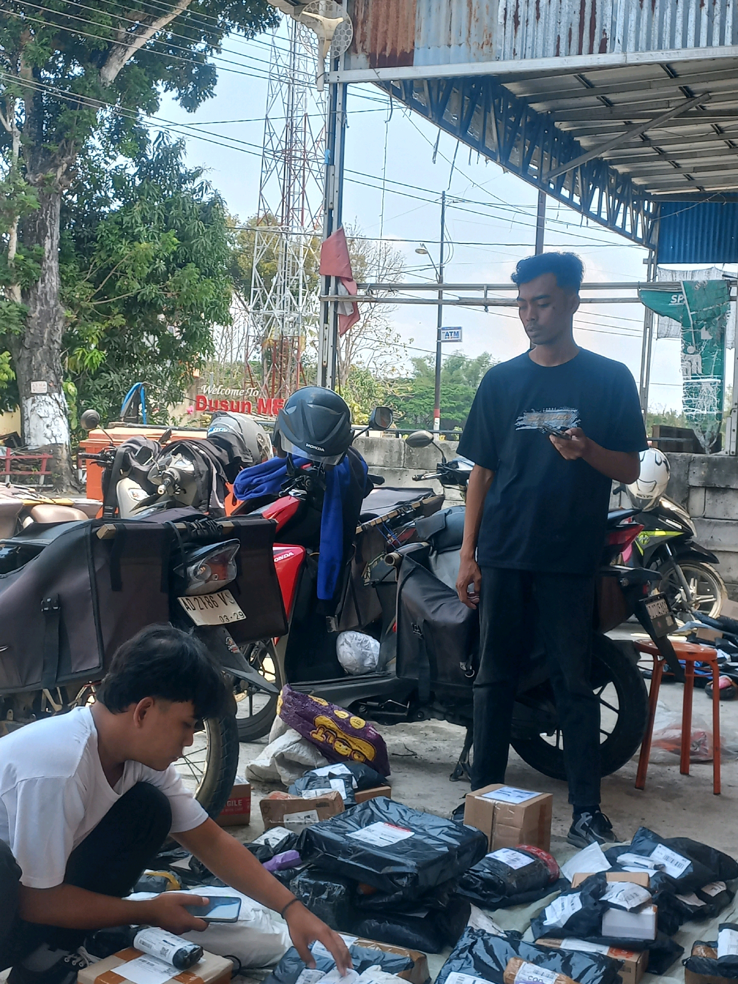 In Baturetno, Indonesia, a scene unfolds with two men amidst a collection of motorcycles and parcels, possibly in a delivery or sorting area. The environment appears to be outdoors, under a covered structure with a corrugated metal roof, likely near a road given the presence of overhead wires and a distant transmission tower.
One man, wearing a white t-shirt and black pants, is crouched down, actively handling and inspecting parcels. He is holding a smartphone in his left hand and reaching out with his right to touch a package. His focus is on the task at hand, surrounded by numerous black plastic-wrapped packages and some cardboard boxes.
The second man, dressed in a black t-shirt and black pants, stands a few feet away, looking down at his own smartphone. He appears to be in a more passive stance, observing or engaged with his device.
Several motorcycles are parked in the vicinity, some equipped with large saddlebags, suggesting their use for transportation or delivery. Helmets are visible on the motorcycles and one is resting on the ground. Various items, including blue fabric and other unidentified objects, are also present. The ground is paved and shows signs of being an active workspace. The overall impression is one of a bustling logistical operation, possibly a courier service or a small business involved in shipping goods.