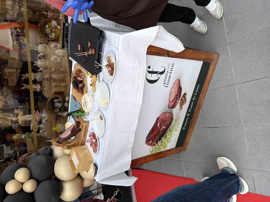 An outdoor food sampling station is positioned on a grey tiled pavement in front of a retail storefront in Felgueiras, Portugal. A rectangular table, covered with a white tablecloth, holds various food items. On the left, a whole cured ham (likely *presunto*) is mounted on a wooden stand, with thinly sliced pieces presented on a small white plate below it. Adjacent is a wooden box containing white napkins. In the center, several rounds of cheese, various types of cured sausages (some whole, some sliced), and a plate of bread or crackers are displayed. A small green bottle is visible behind these items. On the right, a black electric griddle is cooking small pieces of sausage, some with toothpicks inserted, and other cooked pieces are on small white plates.
Behind the table, the storefront window displays numerous decorative items, including Easter bunny figurines, teapots, and ceramic eggs, suggesting a gift or gourmet shop. A large arrangement of black and beige balloons is partially visible on the left, bordering the storefront. Beneath the table, a framed advertising sign depicts various cured meats with herbs, bearing the text "Quinta dos Fumeiros" and "Poiares Ponte de Lima."
The lower body of two individuals is visible. On the left, a person wears blue jeans and light-colored sneakers, holding dark sunglasses. A red floor mat is partially visible under their feet. On the right, a person wears a brown apron over dark trousers and white/gold sneakers; their right hand, clad in a blue disposable glove, is positioned near the electric griddle.