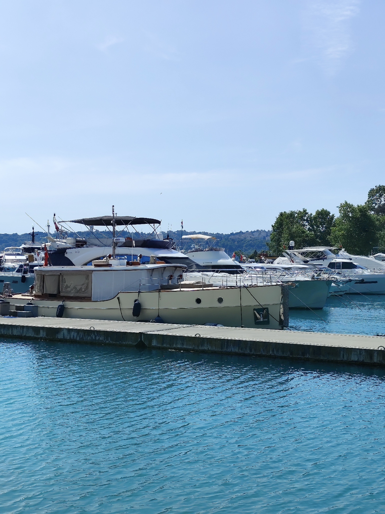 The photograph captures a marina scene in Sarıyer, Turkey, featuring several boats docked alongside a concrete pier. The water is a bright turquoise, exhibiting gentle ripples across its surface. A variety of yachts are visible, with one boat in the foreground positioned against the pier, displaying a light tan or beige hull with a prominent window. Other boats are docked behind this one, including a larger, white motor yacht with a sunshade. The background reveals trees on the distant shore, possibly indicating a wooded area or coastline. The sky is a clear, light blue, suggesting a bright, sunny day.