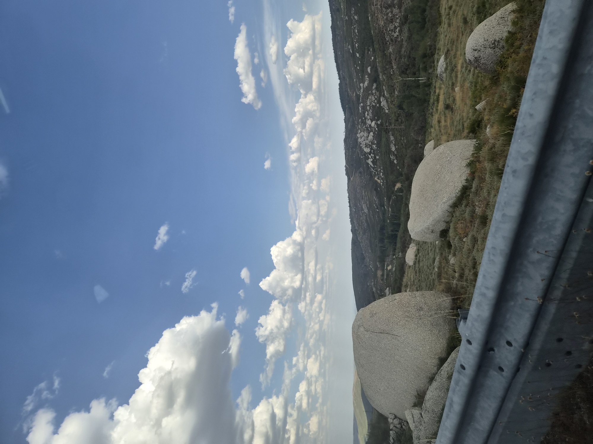 A landscape image captures a vast expanse of rolling hills under a bright blue sky dotted with fluffy white clouds. In the foreground, a section of a metallic guardrail is visible, with dry grass and small bushes growing around it. Scattered across the middle ground are several large, smooth, grey boulders, some appearing partially embedded in the earth. The hills in the distance are covered in a mix of vegetation and smaller rocks, with a few scattered trees. The overall scene suggests a natural, undeveloped area, possibly on the outskirts of Gouveia, Portugal.