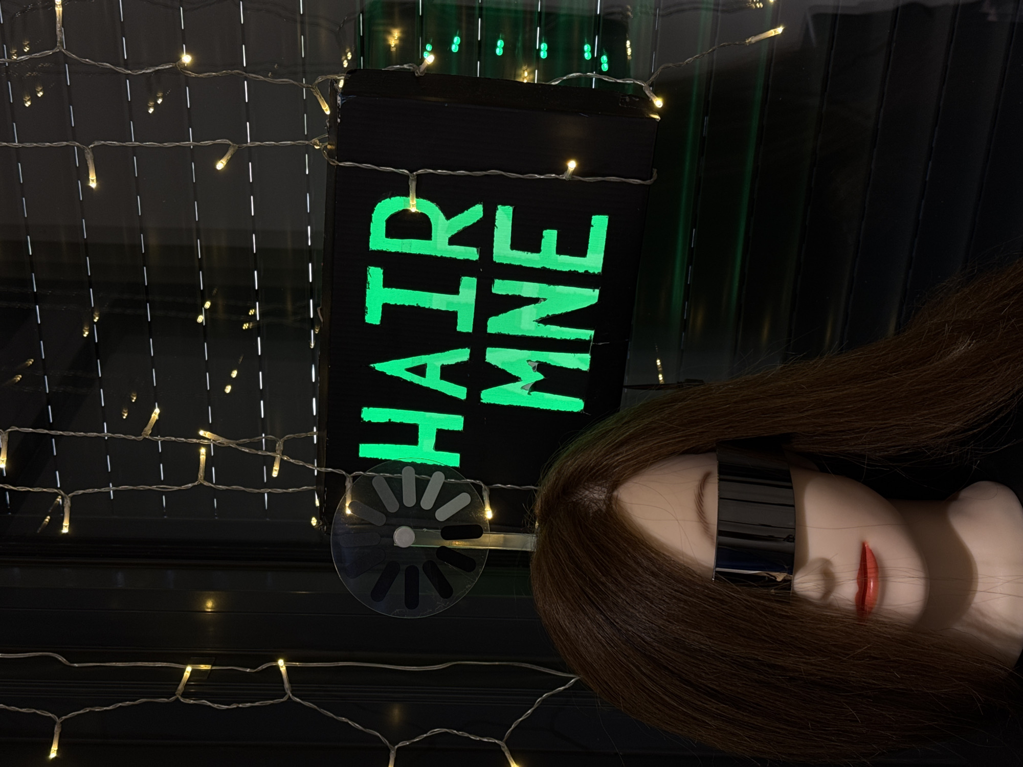 A mannequin head with brown hair and sunglasses is next to a sign that says "HAIR MNE" in green neon letters. The scene is decorated with strings of small, warm-toned fairy lights. The location appears to be a commercial area, based on the signage, which suggests a business related to hair care or styling. I lack the information necessary to confirm that this image was taken in Budva, Montenegro.