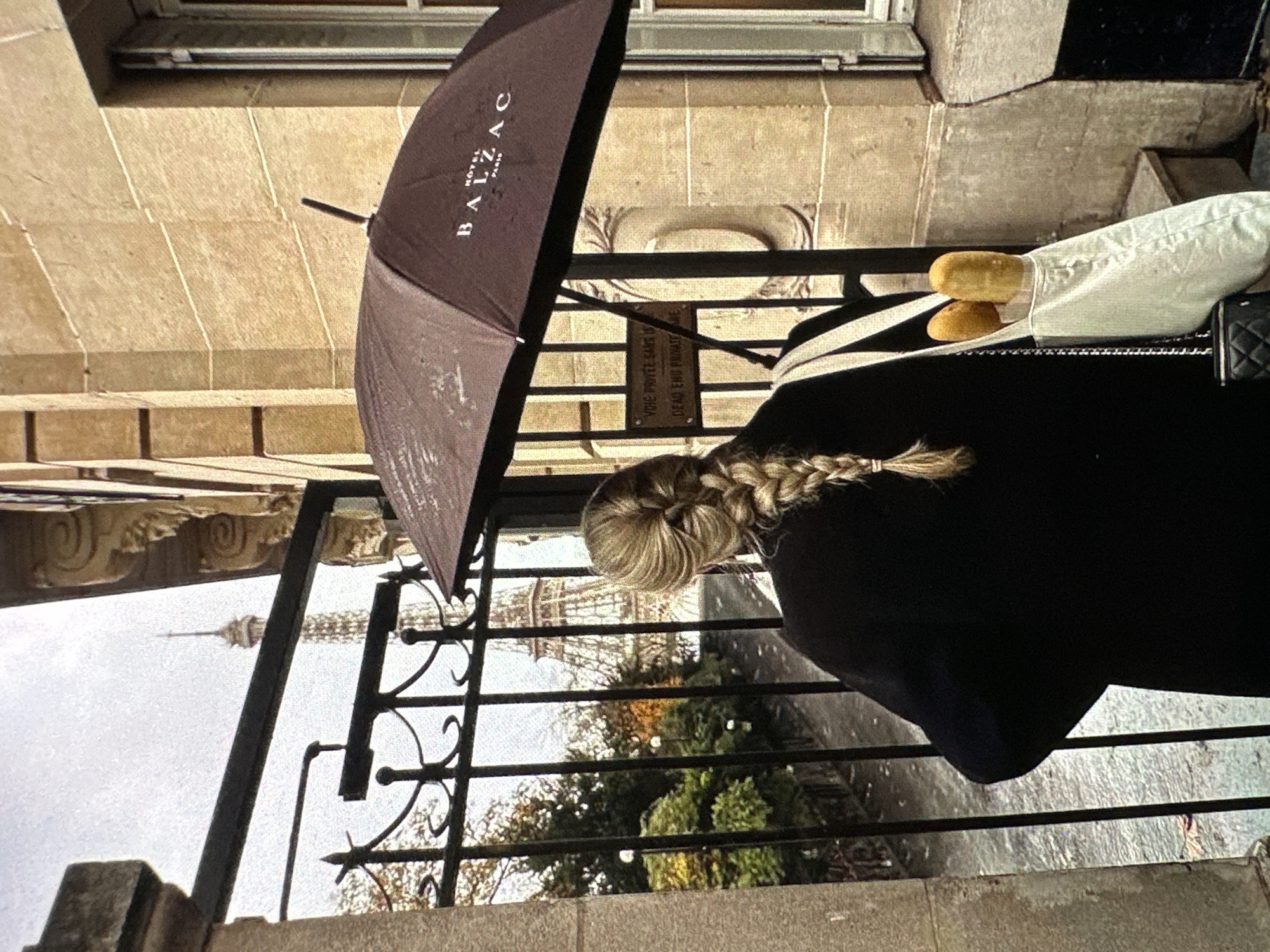 A medium shot shows a woman facing away from the viewer, with a braided hairstyle, wearing a black coat, standing near a wrought-iron railing. An open umbrella with "HOTEL BALZAC PARIS" printed on it is positioned to the left, obscuring part of the background. Two baguettes are in a white cloth bag with straps. The Eiffel Tower is visible through the wrought iron. The location is Paris, France, not Yerevan, Armenia. The scene appears to be taken from an elevated perspective.