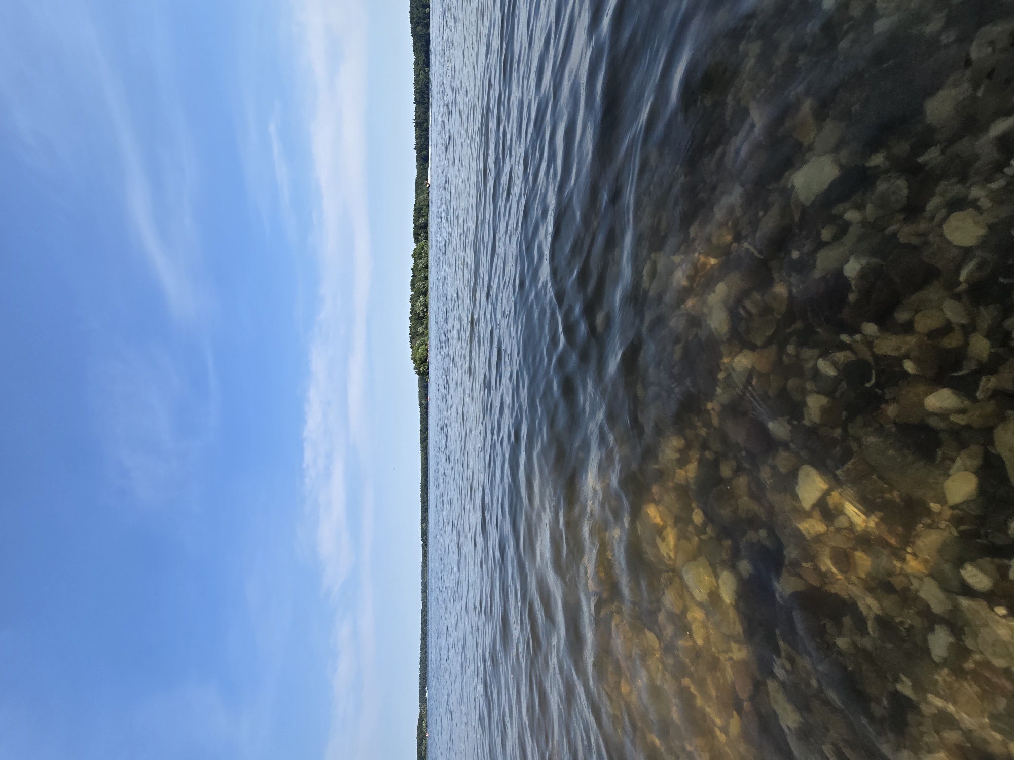 The image depicts a serene natural scene of a lake under a clear blue sky with sparse white clouds. In the foreground, the shallow, clear water allows a view of the rocky lakebed, with various-sized stones and pebbles visible. The water surface exhibits gentle ripples that distort the view of the lakebed. In the midground, the lake stretches to the horizon, where a dense line of green trees forms the shoreline of an island or distant landmass. The far background is a hazy, darker line of trees against the horizon. Two small, white objects, possibly boats or jet skis, are visible on the water in the distance. The sky is a prominent feature, occupying the upper half of the frame, with streaks of wispy clouds. The scene appears to be located in or near посёлок гидроузла имени Куйбышева, Russia, given the context.