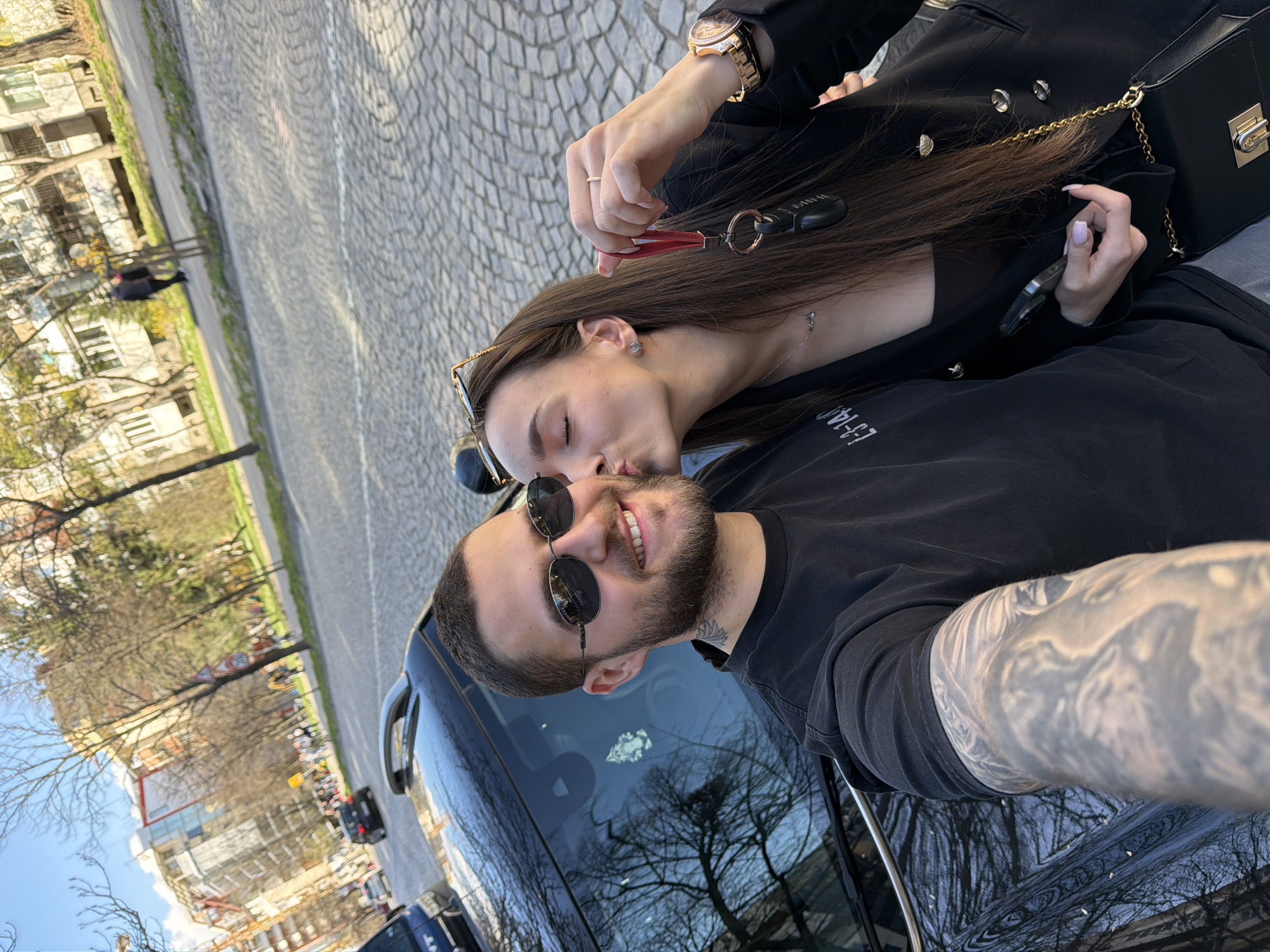 A photograph shows a man and a woman in an embrace. The man is wearing sunglasses and a black shirt. His arm is heavily tattooed. The woman is kissing the man on his cheek. She is wearing a black blazer, a gold watch, and holding car keys. Buildings and trees line the background. The image was taken in Sofia, Bulgaria.