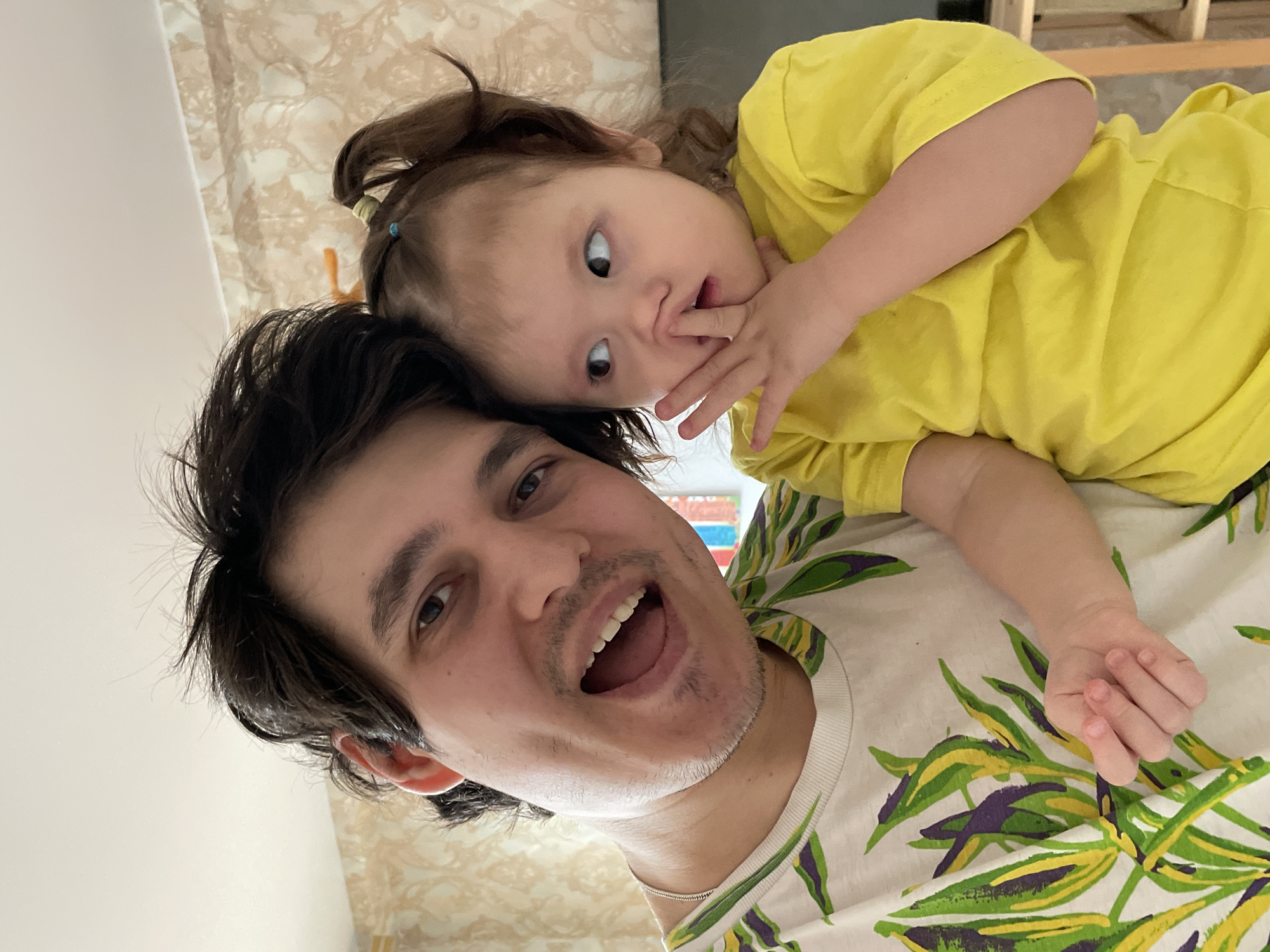 A medium shot depicts a man and a child indoors. The man is lying on his back, smiling with his mouth open, and has dark hair and stubble. He is wearing a white t-shirt with a green and purple leaf pattern. A child is lying on top of him, wearing a yellow t-shirt, and has light skin and dark hair pulled back with a blue hair tie. The child is looking at the camera with their fingers near their mouth. The background is a light-colored wall with a patterned design. There is no evidence in the image to support that it was taken in Izhevsk, Russia.