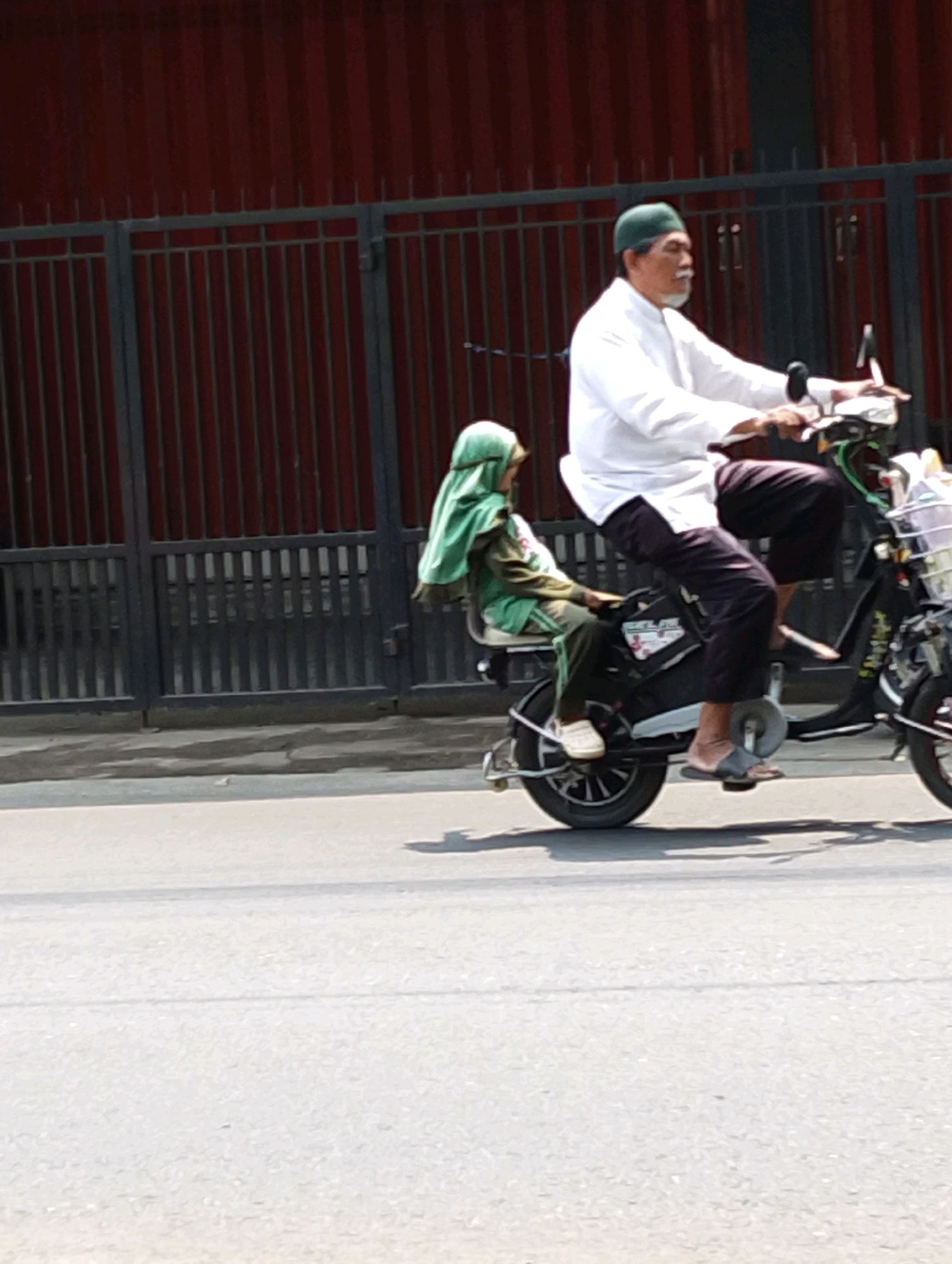 In Gempeng, Indonesia, a man wearing a white long-sleeved shirt and a green cap rides a black electric scooter on a paved road. He has a mustache and a goatee. Behind him, a child wearing a green hijab and a green jacket sits on the scooter. The scooter has a basket on the front and a seat in the back for the child. In the background, a dark red building with vertical lines is visible behind a black metal fence. The scene is lit by sunlight, casting shadows on the road.