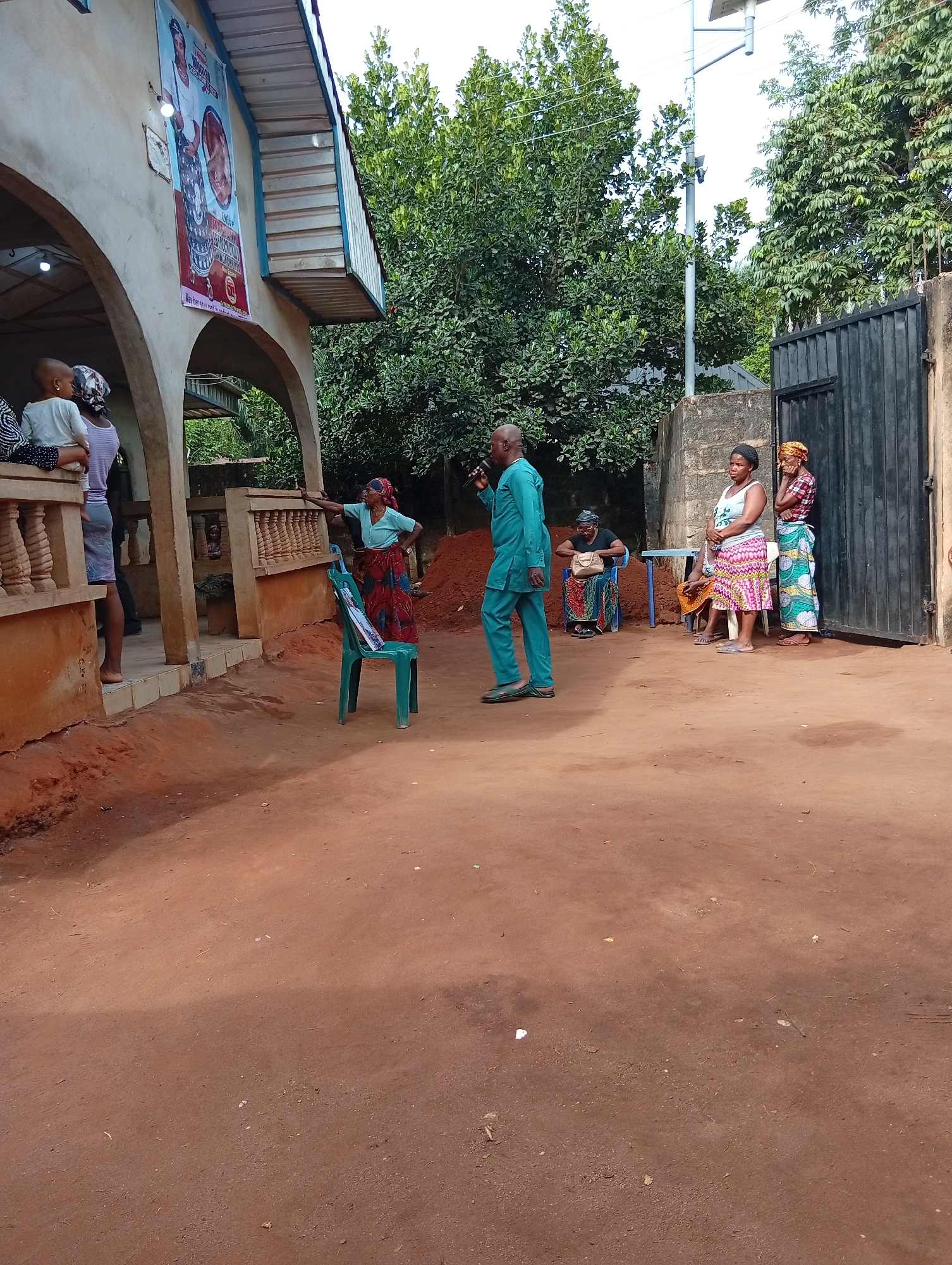 The image shows a gathering of people in an outdoor setting, likely in a residential or community area. The ground is reddish-brown dirt. A man in a teal-colored suit is speaking into a microphone. Several women are standing and listening to him. Some are wearing traditional African attire. One woman is leaning on a green plastic chair. In the background, there is a building with arched openings. A large poster hangs on the building. Beyond the people, there is a gate, a wall, and trees. A streetlight is mounted on a pole. The city is Unknown, Unknown.