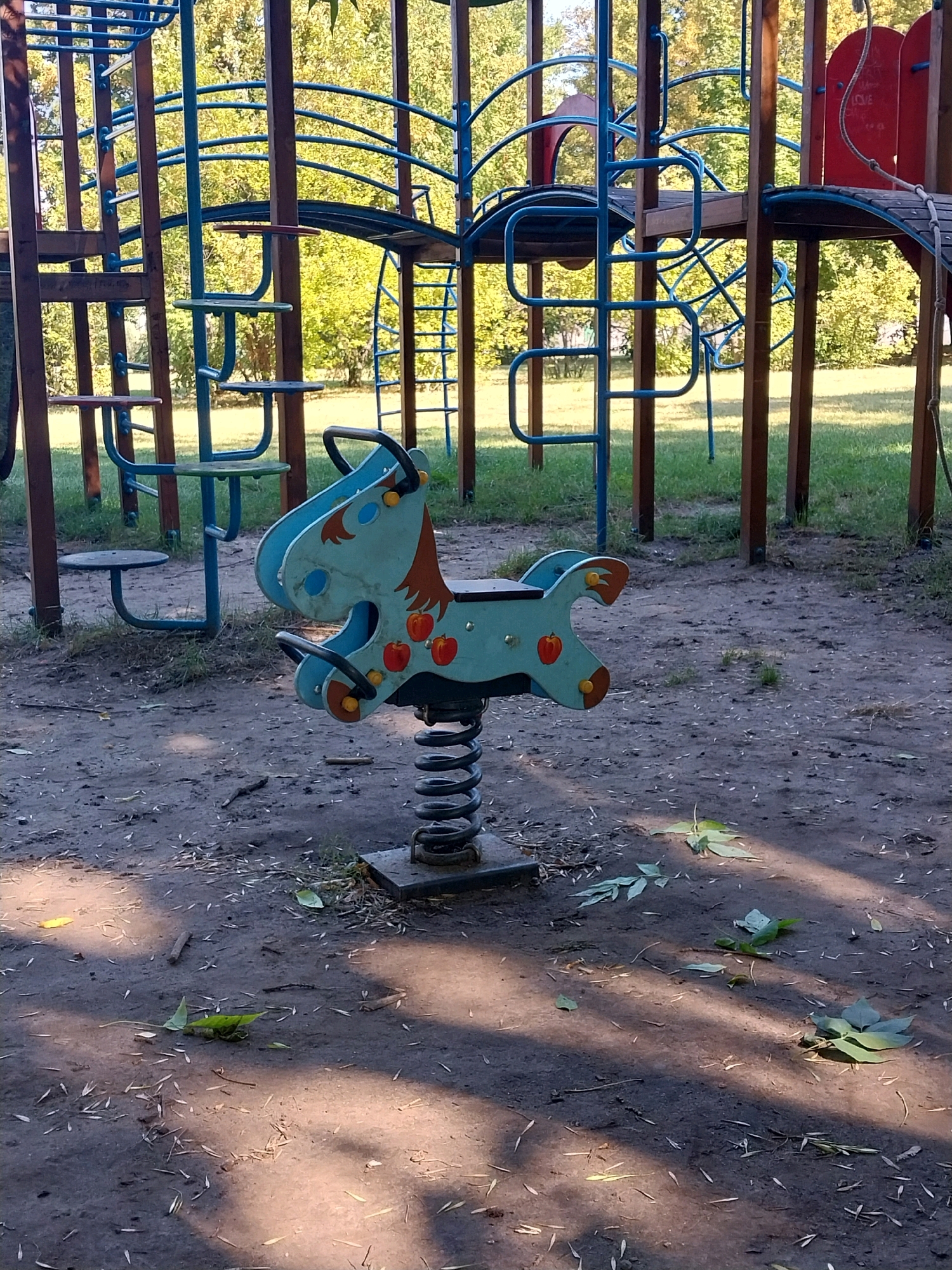 The image depicts a bright, sunny day at a playground, likely in the city of Doslidne, Ukraine. In the foreground, a light blue, spring-mounted toy horse with red apple designs and brown mane and ears is positioned on a patch of dark, sandy ground littered with leaves and twigs. The horse has black handles and yellow bolt heads. Behind the toy horse, a larger, multi-level playground structure made of dark brown wooden posts and blue metal bars dominates the background. This structure includes various platforms, ladders, and archways. Green grass and deciduous trees with yellowing leaves are visible behind the playground equipment, suggesting an autumn season. The overall lighting indicates natural daylight, with shadows cast on the ground from the playground structure.