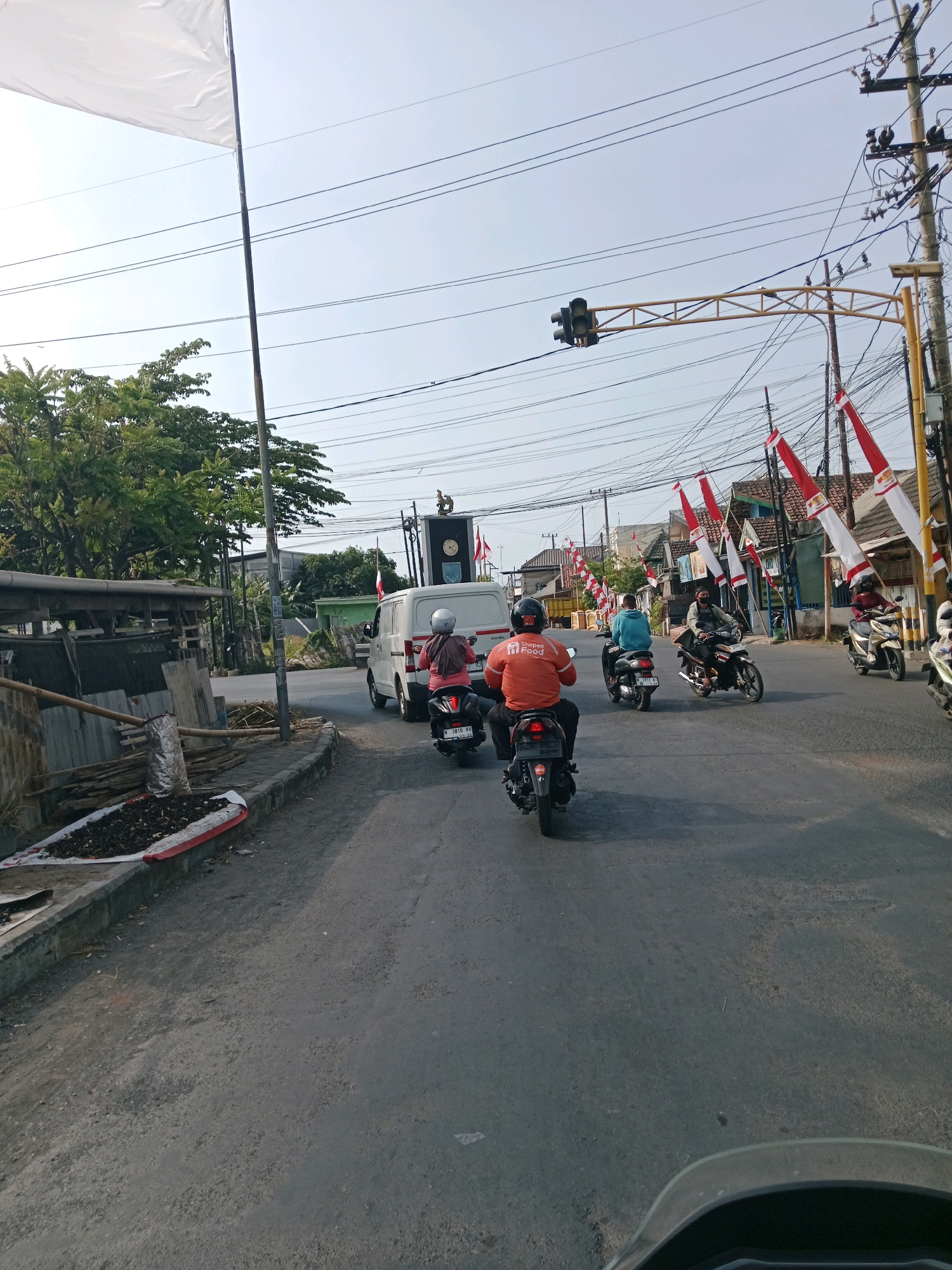 The image captures a street scene in Probolinggo, Indonesia. In the foreground, the perspective is from a vehicle, showing a portion of its windshield and handlebars. The road is paved and appears to be a street with moderate traffic. Several motorcycles are visible, with riders and passengers. One rider in the middle lane wears an orange shirt with the "Shopee Food" logo. A white van is also present.
Along the sides of the road, there are some structures. On the left, a simple roadside stall or shelter is visible, with some materials stacked next to it. On the right, utility poles with numerous wires are prominent, and red and white Indonesian flags are strung across the street, suggesting a national celebration or event. A traffic light hangs over the intersection. The sky is clear and bright, indicating daytime. The overall impression is a typical urban or suburban street environment in Indonesia.