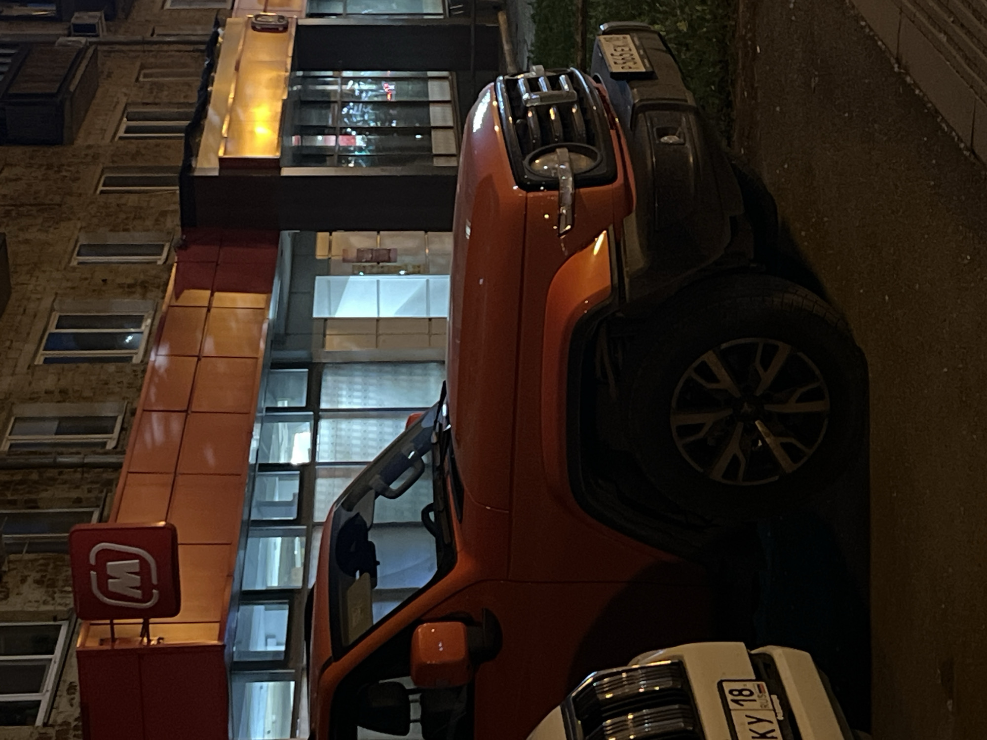 The image captures an urban street scene at night in Izhevsk, Russia, with several parked vehicles alongside a multi-story building. The main subject is an orange SUV, resembling a Jeep Renegade, positioned prominently in the foreground. It features a distinctive front grille with vertical slats, circular headlights, and roof racks.
Behind the orange SUV, a dark-colored car is partially visible, displaying a Russian license plate that reads "P 669 EX 18 RUS." The "18 RUS" regional code indicates the Udmurt Republic, where Izhevsk is located. In the lower right of the image, the front of a white car is visible, also with a Russian license plate showing "KY 18 RUS" and the word "RUS" below.
The building on the left side of the frame features a modern facade, predominantly in shades of orange and dark red. It has numerous rectangular windows, some reflecting the artificial lights from the street or nearby buildings. A prominent red square sign with a white stylized 'M' logo is affixed to the building's facade. This 'M' logo is consistent with the branding of the Magnit (Магнит) supermarket chain, a common retail presence in Russia. The building's architecture suggests a commercial establishment on the lower floors, with potential residential or office spaces above.
The scene is lit by artificial light sources, indicating it is nighttime. No people are visible in the frame. The overall impression is one of a typical urban evening, with vehicles parked near a commercial building.