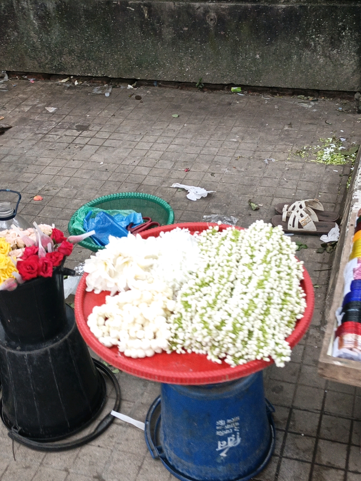 An outdoor scene in Dhaka, Bangladesh, depicts a street vendor's stall. A variety of flowers, primarily white jasmine garlands and red roses, are displayed in black and red containers. These containers are placed on top of blue and black buckets. In the background, a green basket holds plastic bags. The ground is paved with square tiles, and debris is scattered around the area. A pair of sandals rests on the pavement near a wooden stall displaying colorful coiled items. The overall environment suggests a busy urban setting.