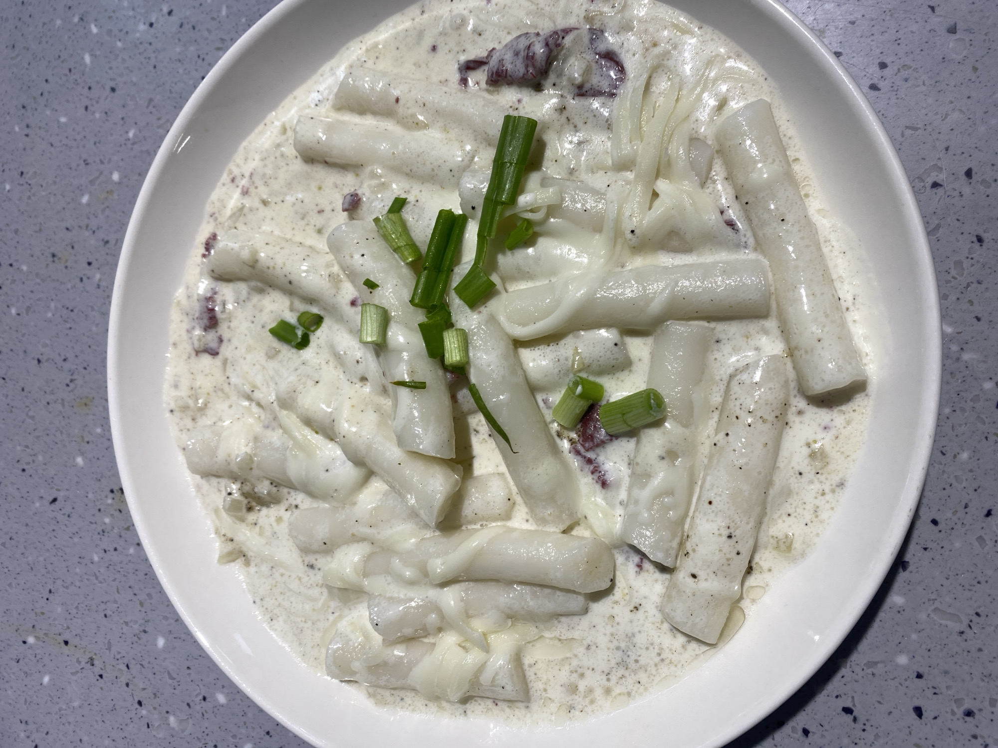 A white ceramic bowl is filled with creamy tteokbokki. The tteokbokki consists of cylindrical rice cakes, some of which are slightly flattened and twisted. They are coated in a rich, creamy white sauce speckled with black pepper and some small, reddish pieces which appear to be diced red onion or possibly bacon. Scattered throughout the dish are thin strands of melted mozzarella cheese, giving it a stringy appearance. A few chopped green onions are sprinkled on top as a garnish. The bowl is placed on a speckled gray countertop. There are no people or animals visible in the image.