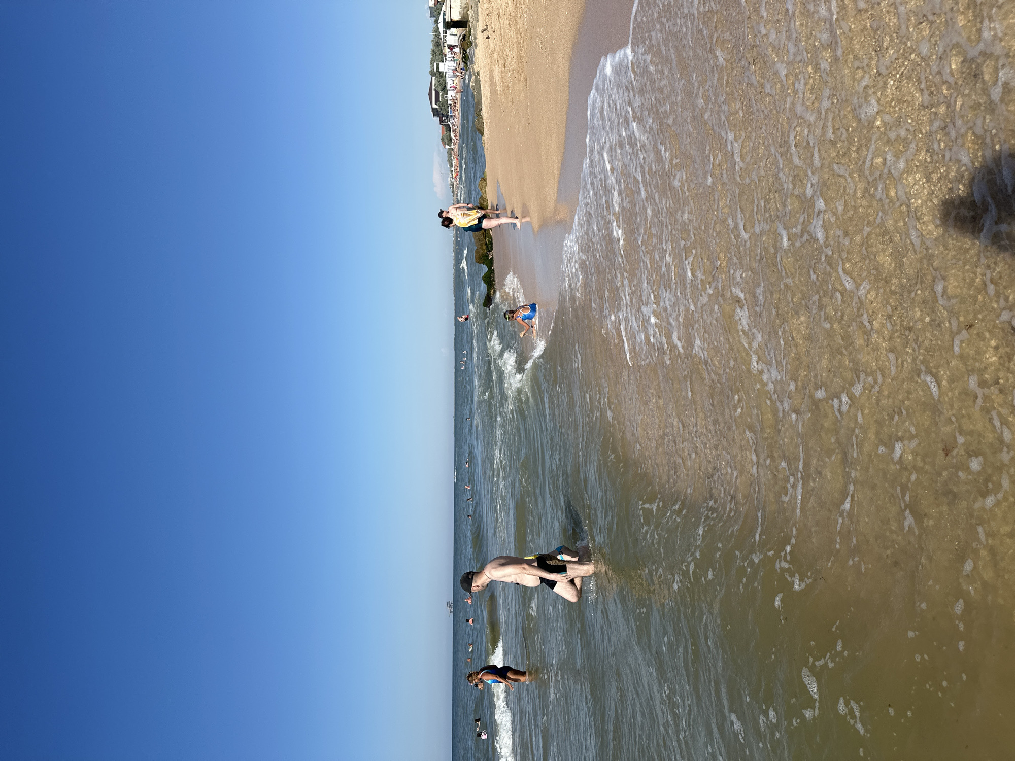The image shows a beach scene in Golubitskaya, Russia. The sky is a clear, bright blue, suggesting a sunny day, possibly late morning or early afternoon. The foreground is dominated by shallow, murky water with gentle waves lapping at the sandy shore. White foam from the waves creates interesting patterns on the sand.
Several people are visible in and around the water. In the mid-ground, a man wearing black shorts and a cap is standing in the water, facing towards the shore. Further out in the water, several other individuals are swimming or wading. Closer to the shore, a woman in a blue swimsuit is partially submerged in the water, looking down.
The beach itself is sandy and appears to have some rocky areas or breakwaters extending into the sea. In the background, along the edge of the sand, there are structures that resemble beachside buildings or kiosks, and a line of trees. More people are scattered along the shoreline and on the sand, indicating it's a popular spot. The overall atmosphere seems relaxed and typical of a summer day at the beach. There is no visible text in the image.